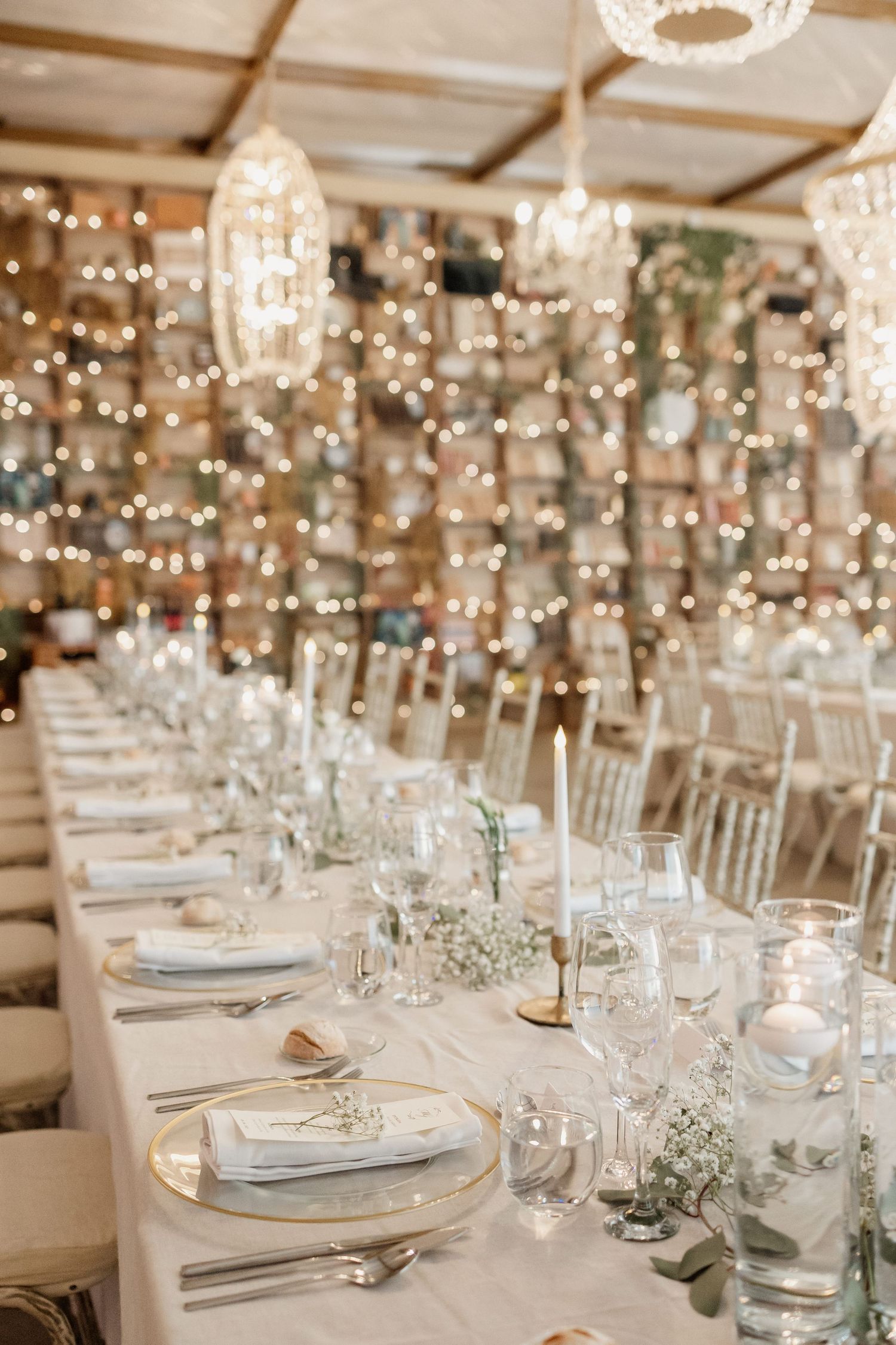 Elegant wedding reception setup with crystal chandeliers, white tablescapes and baby's breath centerpieces under string lights.
