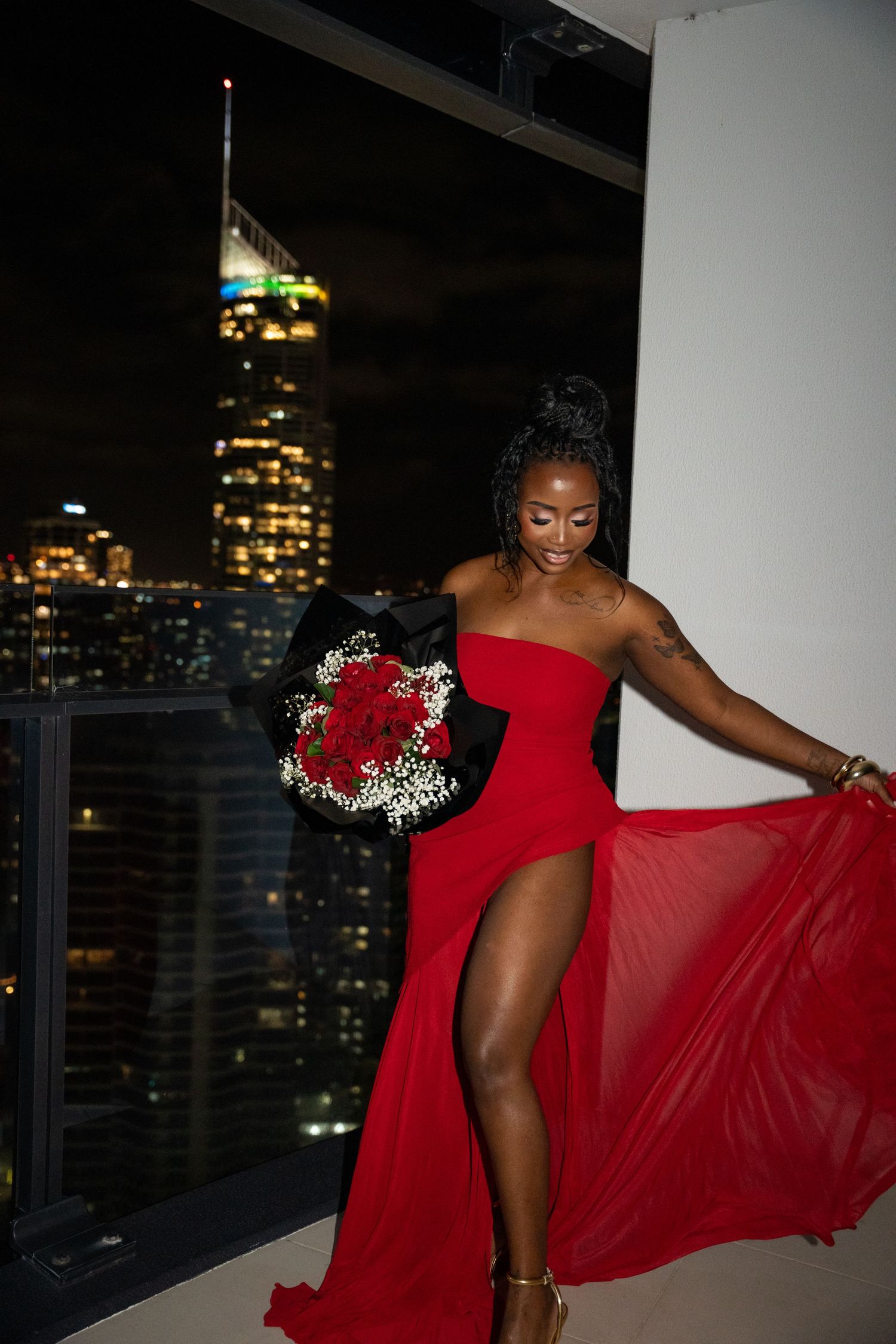 Woman in red strapless gown holding red rose bouquet on high-rise balcony with city skyline at night.