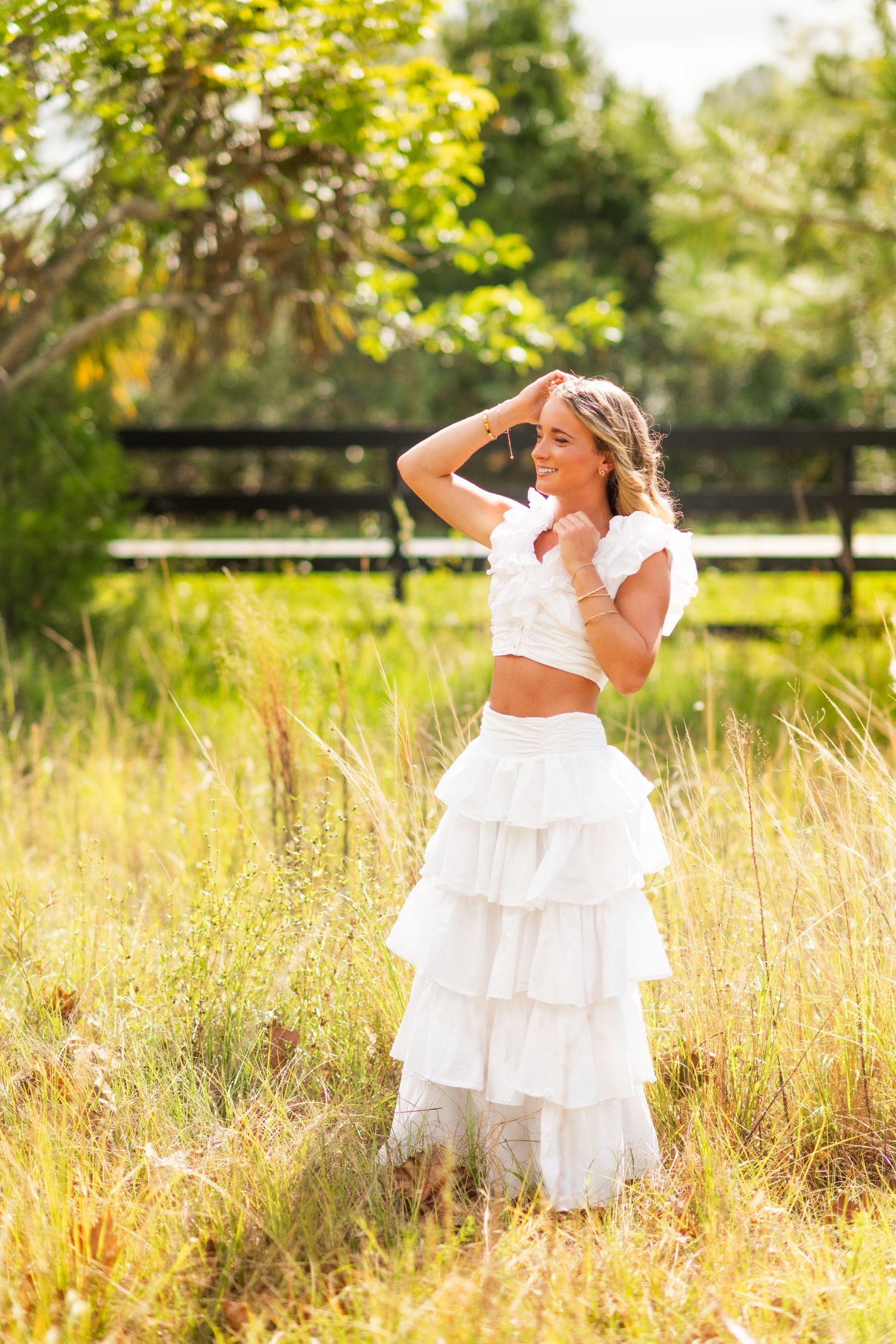 Young woman in white ruffled two-piece outfit posing in golden sunlit field near wooden fence.