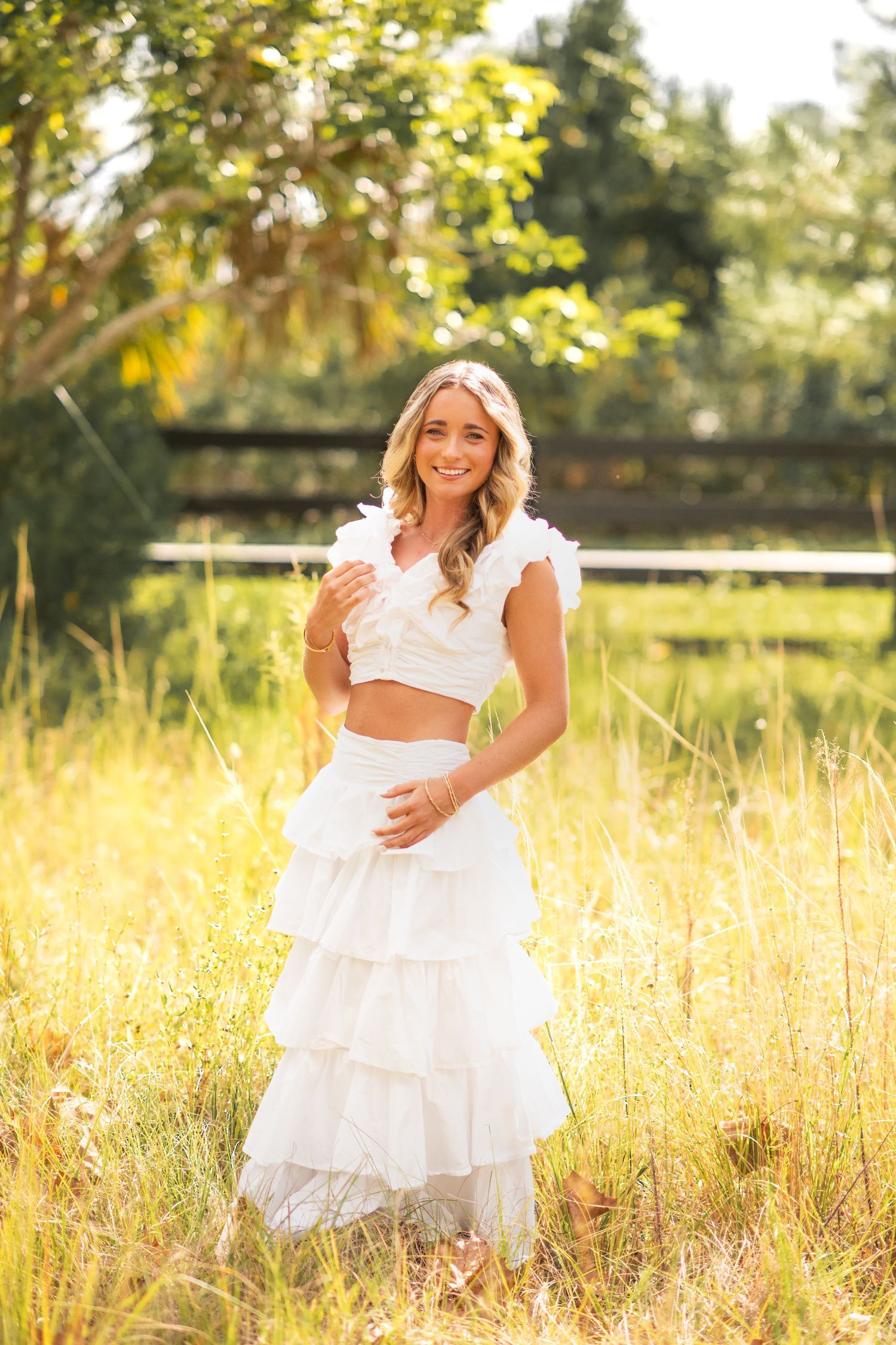 Smiling young woman in white ruffled crop top and tiered maxi skirt standing in a sunny golden field.
