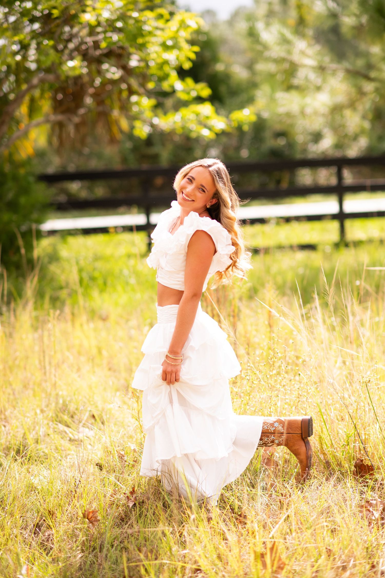 Young woman in white ruffled outfit and cowboy boots walking through a sunny golden meadow.