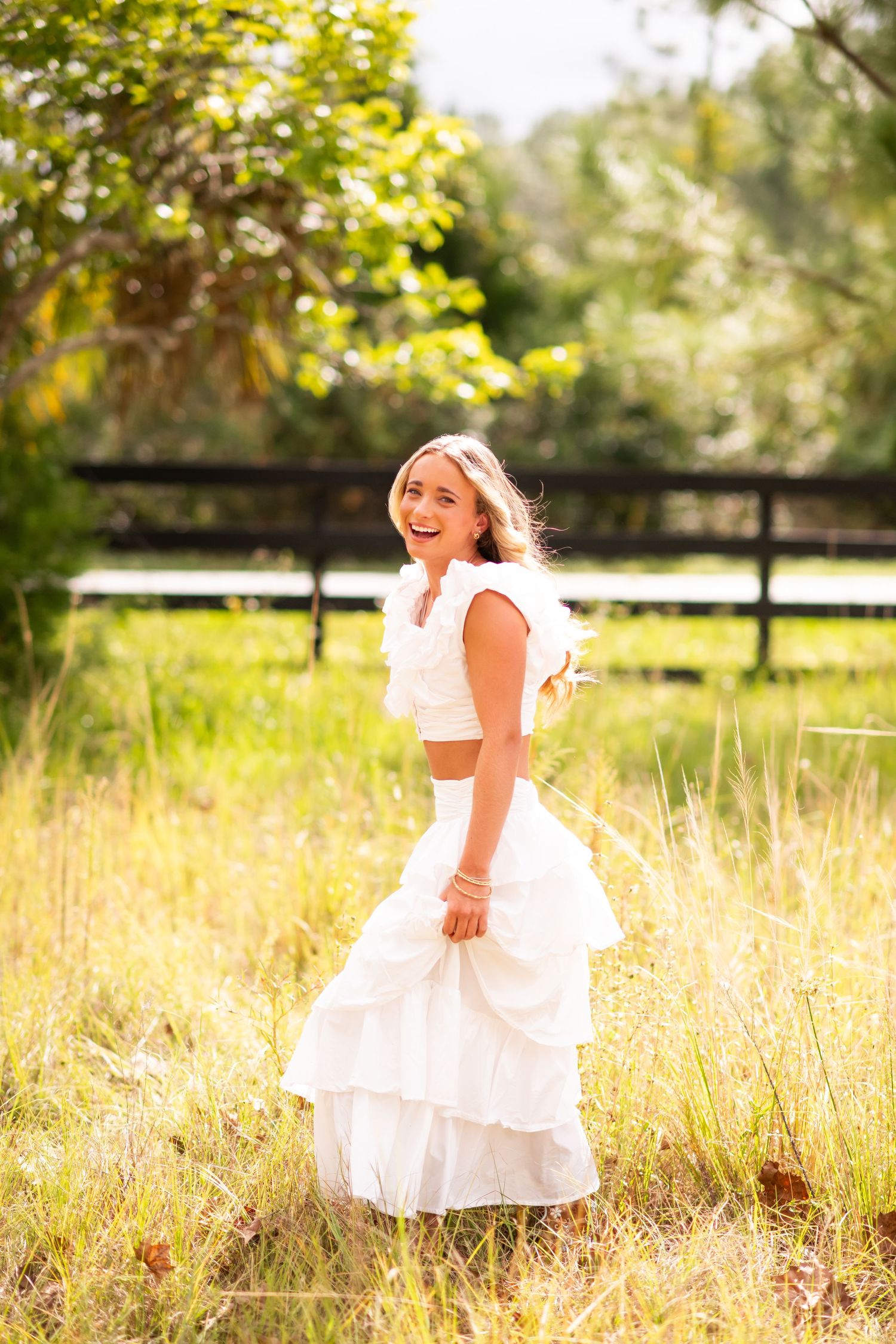 Laughing young woman in white ruffled crop top and skirt standing in a sunny golden field outdoors.