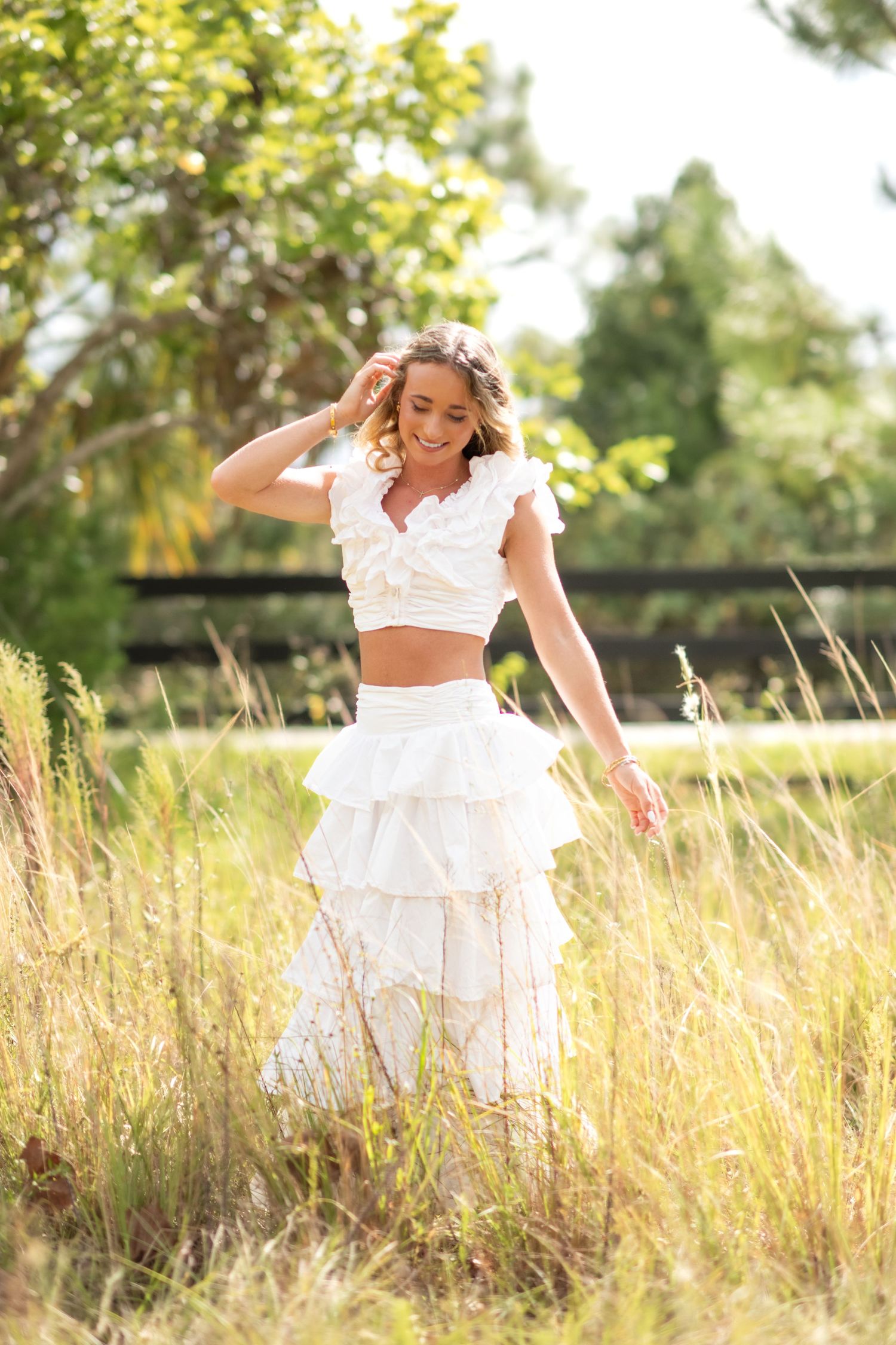 Young woman in white ruffled crop top and maxi skirt smiling in a sunlit field with tall grass.