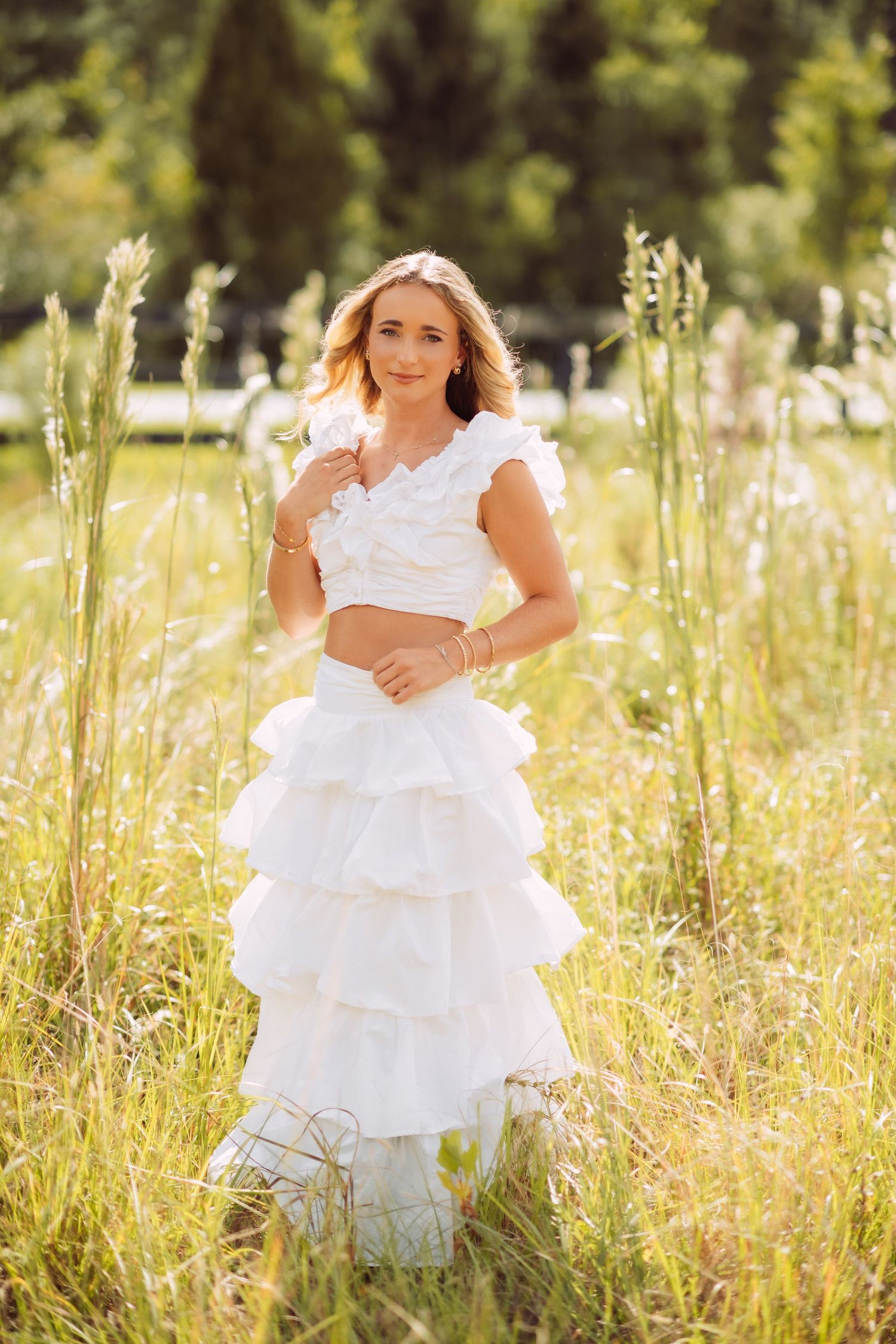 Young woman in white ruffled crop top and tiered skirt stands in sunny green meadow field.