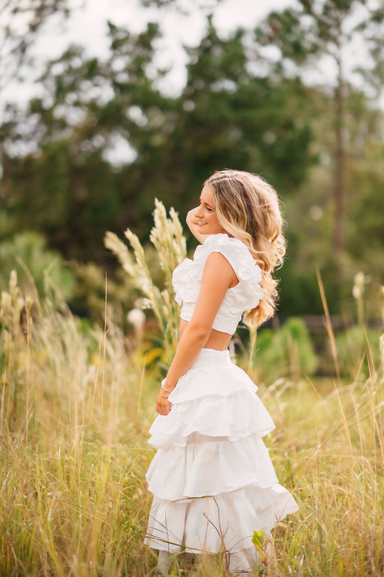 Young woman in white tiered skirt and crop top smiling over her shoulder in a lush green field.