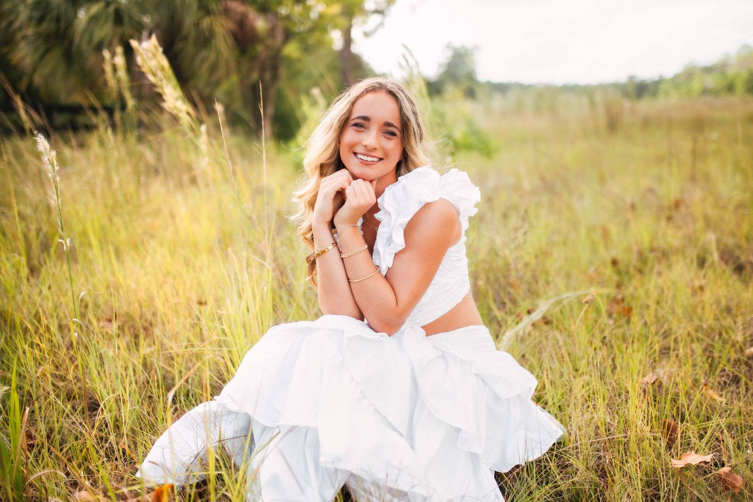 Smiling blonde woman in white ruffled outfit sitting in a sunlit grassy field outdoors.