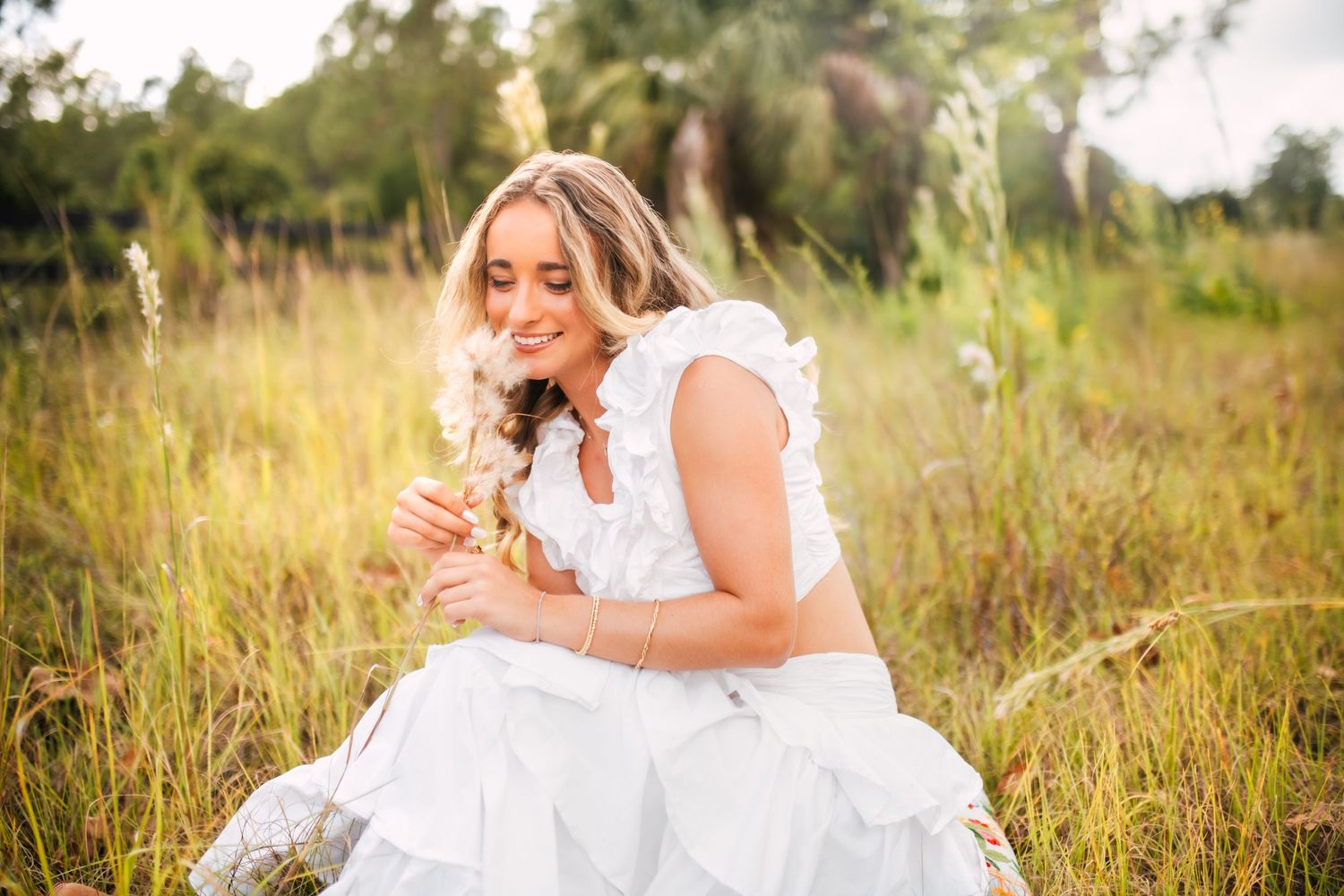 Blonde woman in white dress sitting in golden meadow, smelling wildflowers in soft sunlight.