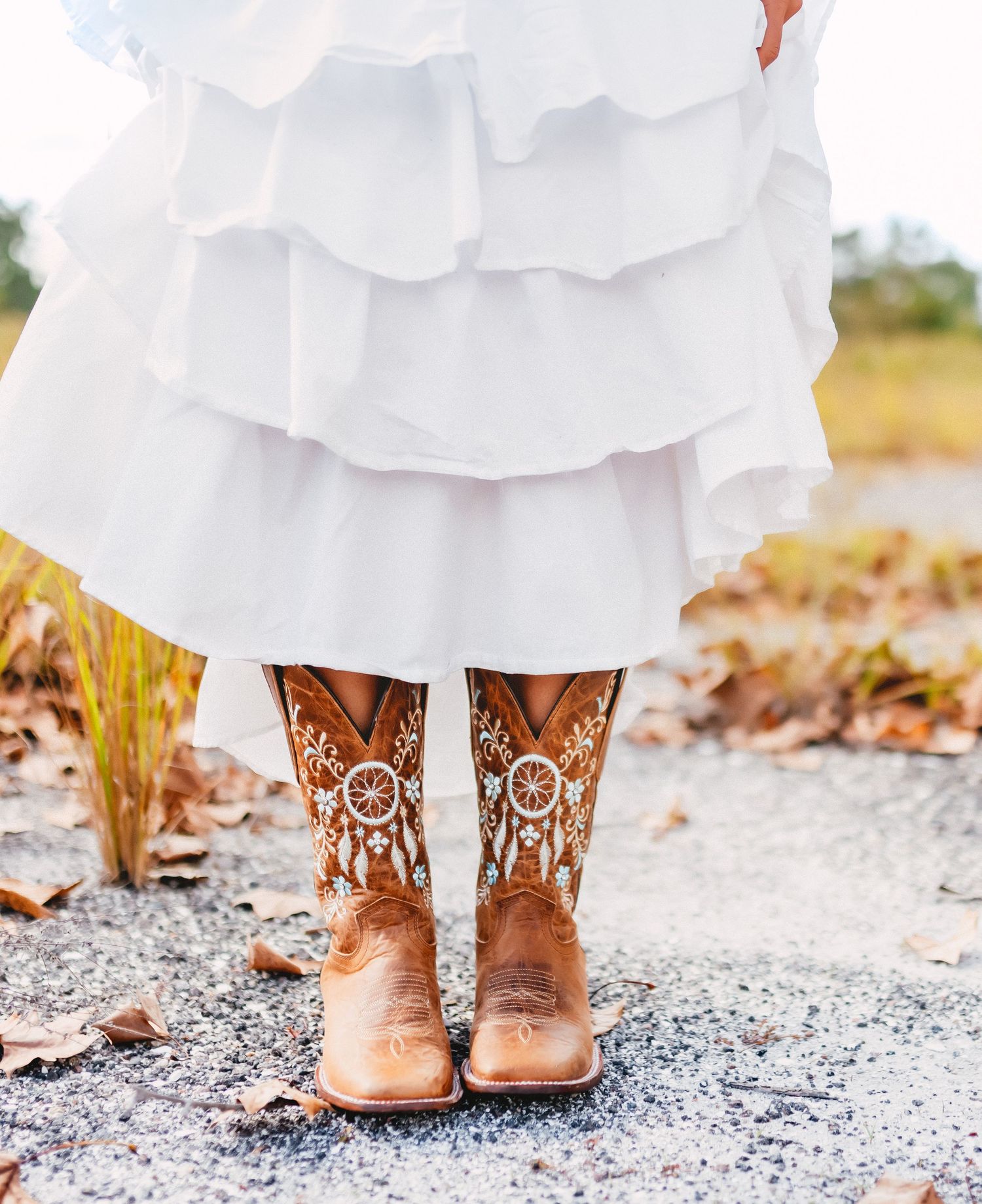 Close-up of decorative brown cowboy boots with dreamcatcher design beneath a white ruffled skirt.