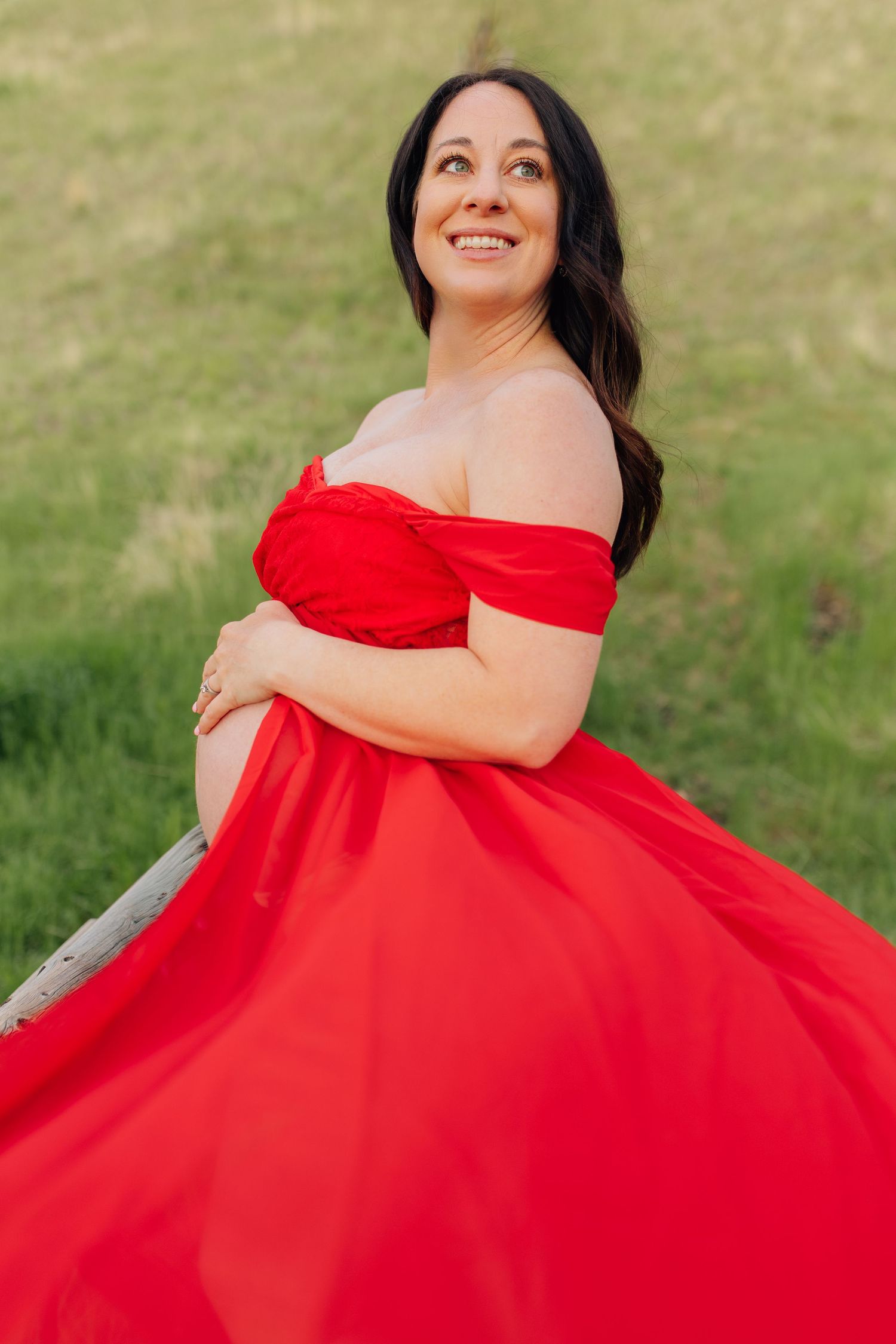 Pregnant woman in flowing red off-shoulder gown smiling outdoors on green hillside.