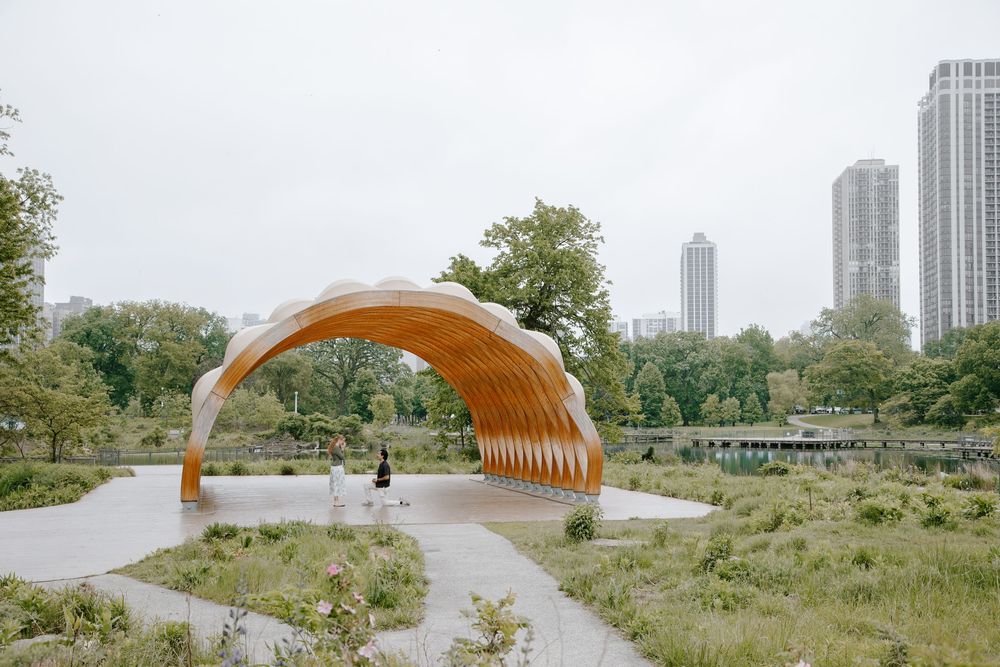A wooden arch sculpture in a park with city skyline, surrounded by greenery and pathways on an overcast day.