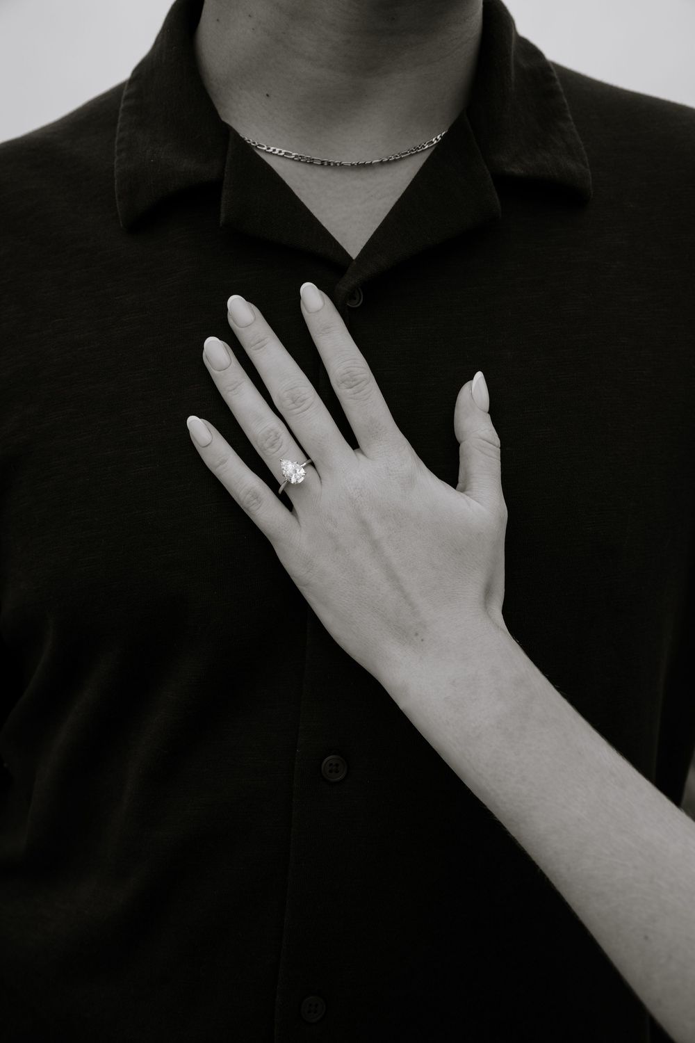 Elegant hand with long nails wearing a floral diamond ring resting on a dark button-up shirt with a delicate chain necklace.