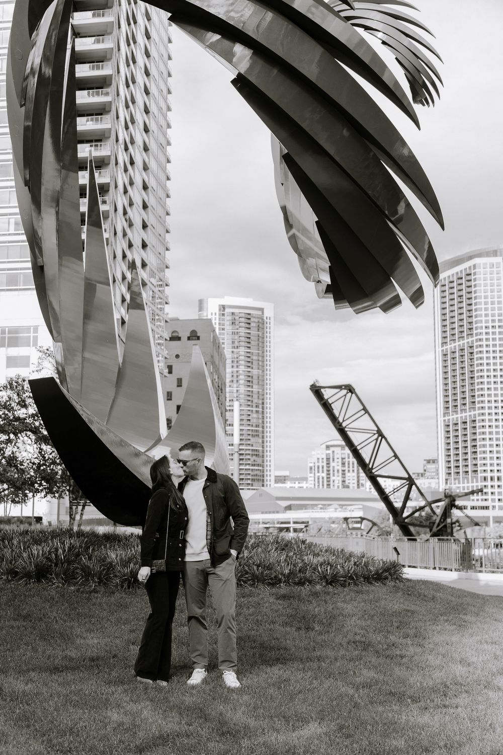 Black and white photo of a couple standing together near a large modern sculpture with city skyscrapers behind them.