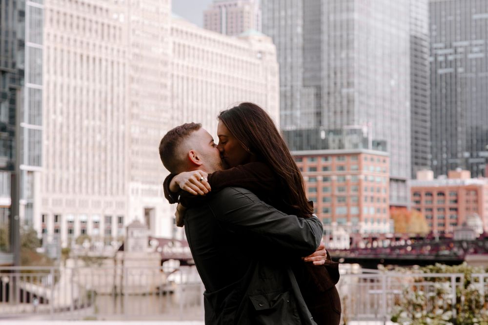 Couple sharing a romantic kiss on a city rooftop with urban skyline and river in the background.