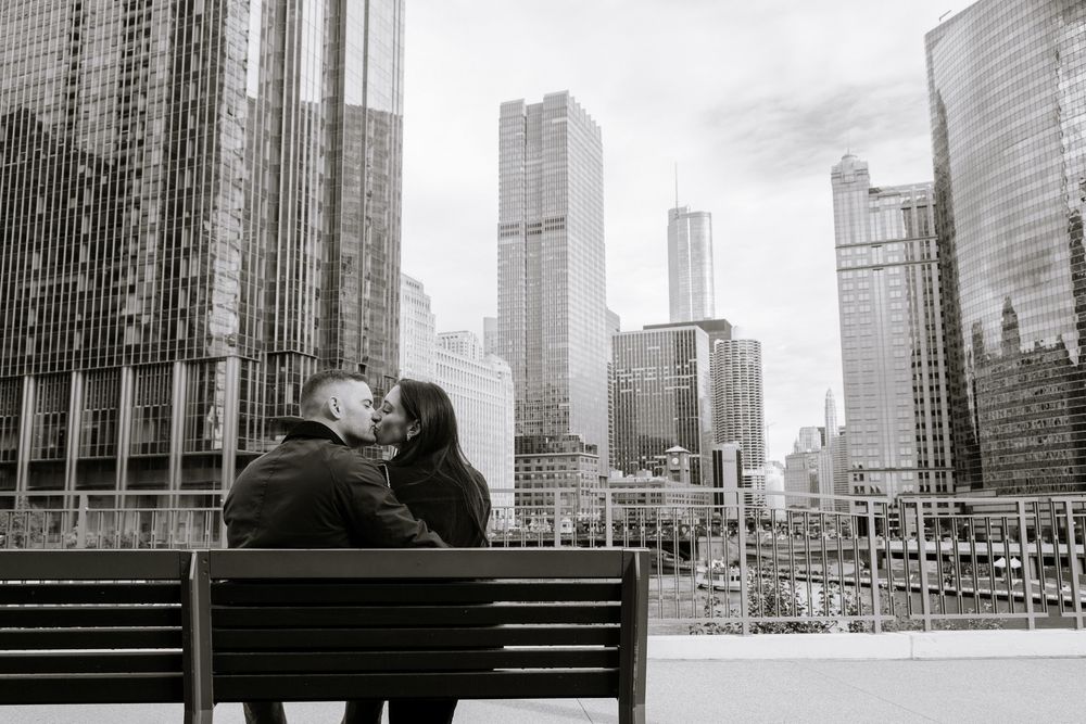 Black and white photo of a couple sharing a kiss on a bench with Chicago city skyline skyscrapers in the background.