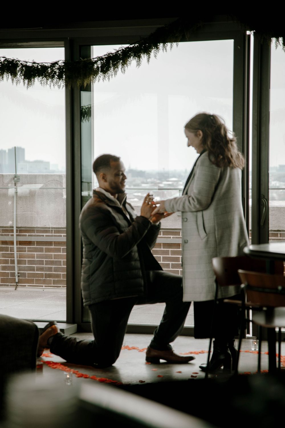 Man proposes on one knee to woman in front of large windows with city views and holiday garland.