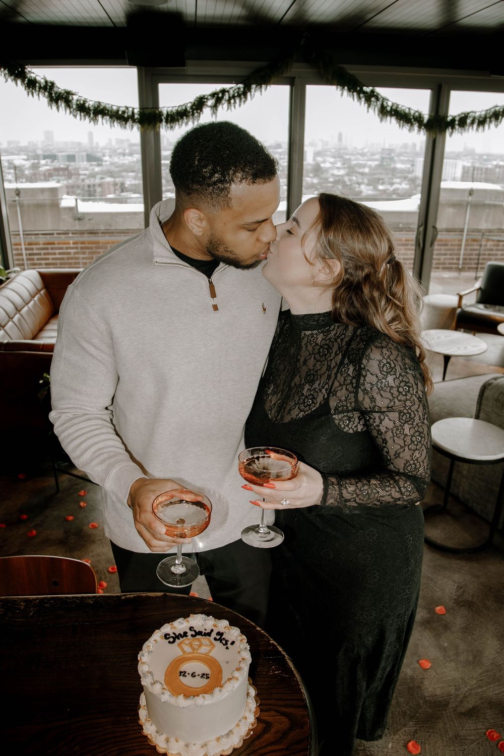 Couple kissing while holding cocktails beside a celebration cake at a rooftop bar with city views.