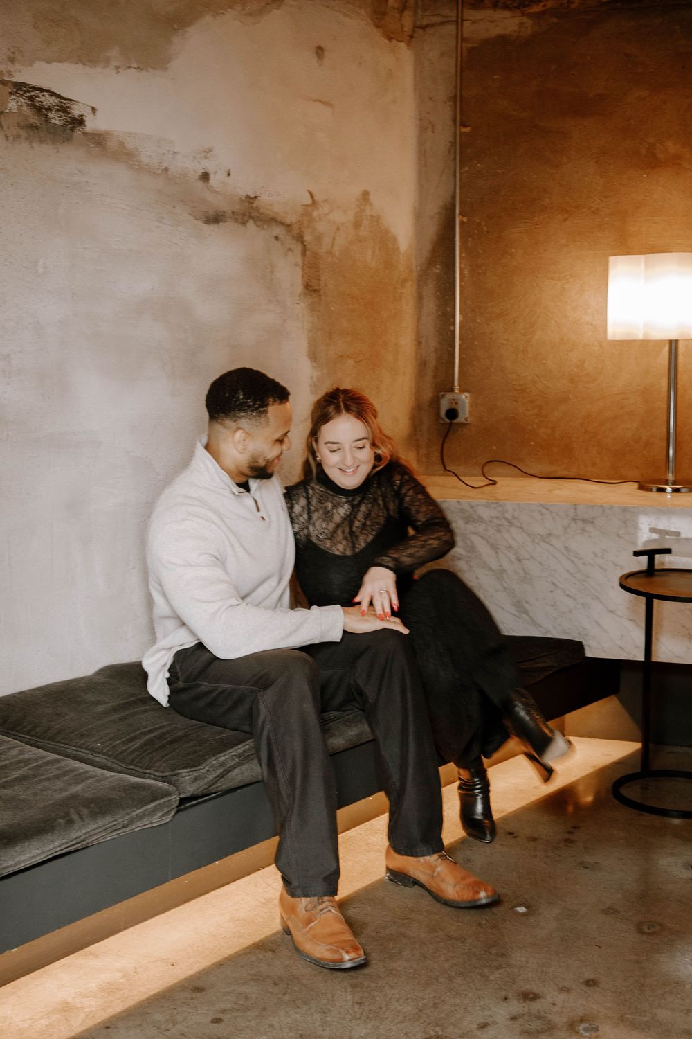 Couple sitting together on a velvet bench in a cozy industrial space, smiling and holding hands.