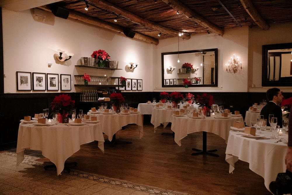 Elegant restaurant dining room with white tablecloths, red poinsettias, wood beam ceiling, and framed artwork on cream walls.
