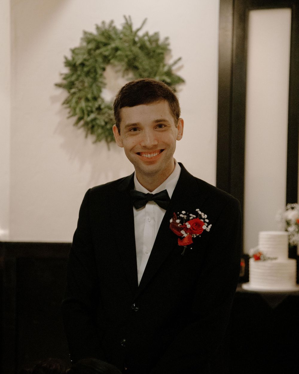 Young man in black tuxedo with red boutonniere smiling at wedding reception with wreath backdrop.