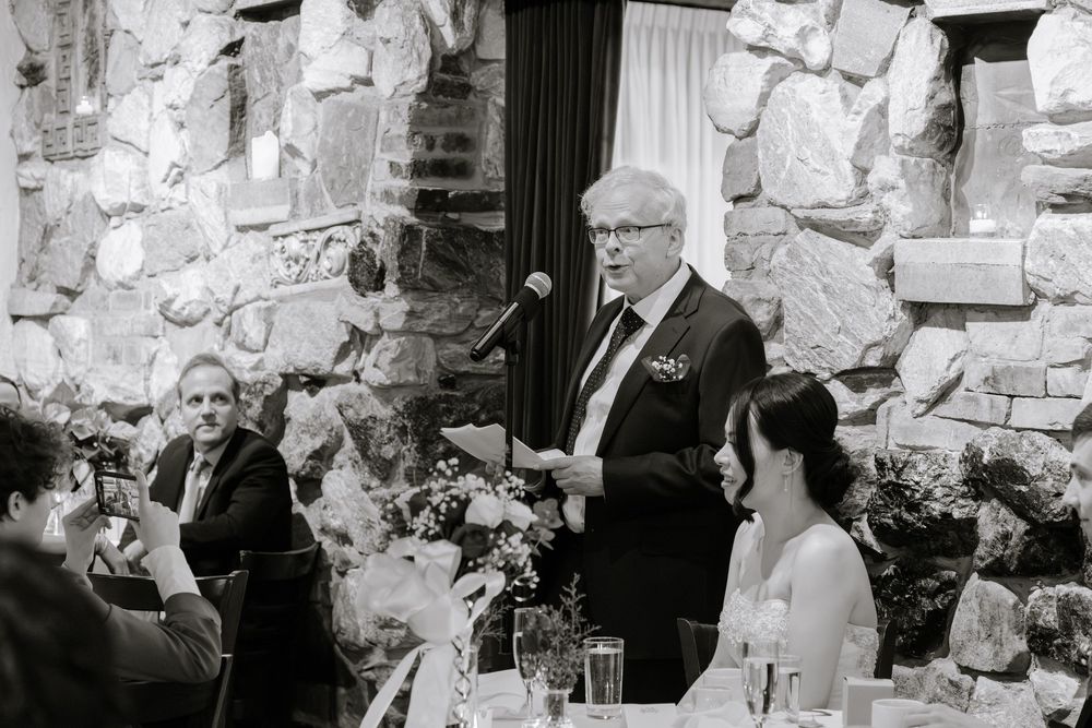Man giving a speech at an elegant wedding reception in a stone-walled venue, guests seated at decorated tables.