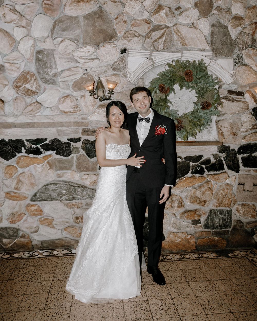 Bride in white gown and groom in black tuxedo pose before a rustic stone wall with a holiday wreath.