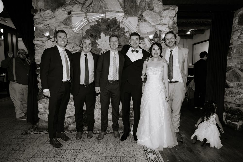 Black and white wedding photo of bride and groom posing with five guests in front of a stone fireplace.