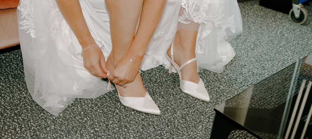 Bride fastening white pointed-toe heels with ankle straps while wearing a flowing wedding dress.