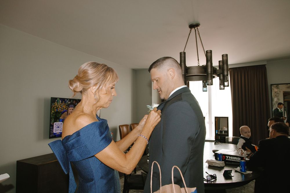 Woman in blue off-shoulder dress adjusting groom's tuxedo bow tie in elegant hotel room before wedding.