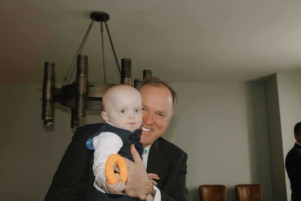Elderly man in suit smiling while holding a happy baby with an orange teething ring indoors.