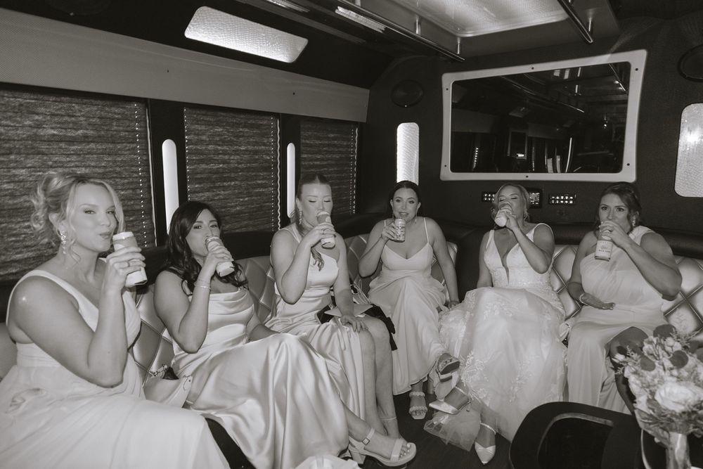 Bridesmaids in white dresses toasting with drinks inside a party bus in black and white photo.