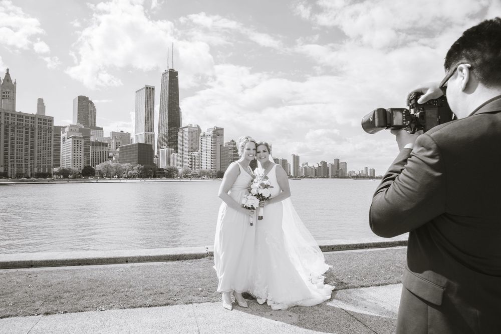 Two brides pose with Chicago skyline backdrop as photographer captures their wedding portrait by the lakefront.