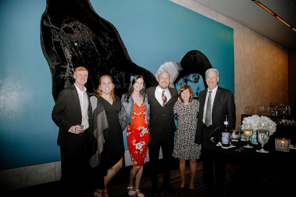 Group of six adults posing together at an elegant evening event with blue wall art backdrop.