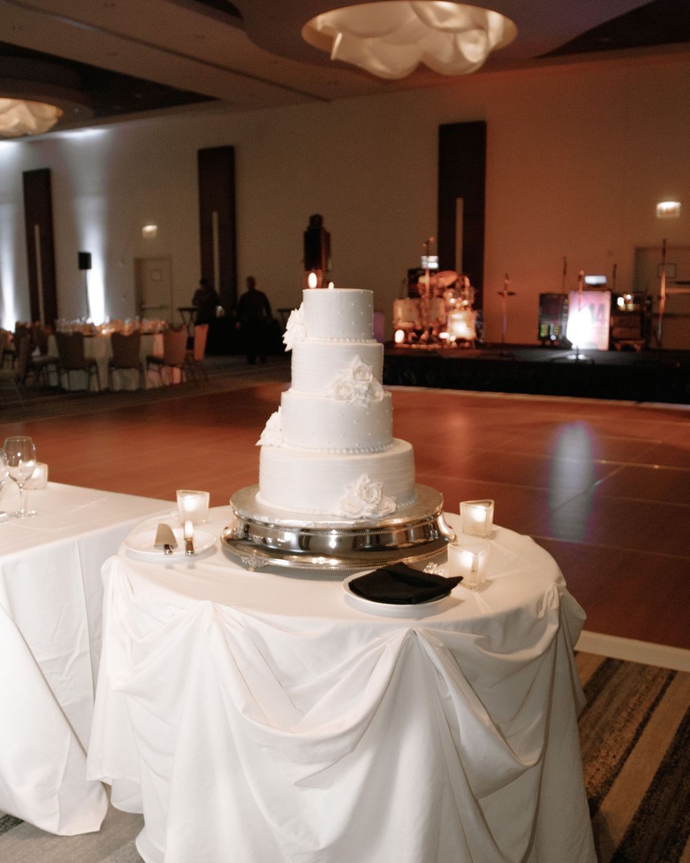 Elegant four-tier white wedding cake on draped table in softly lit ballroom reception venue.