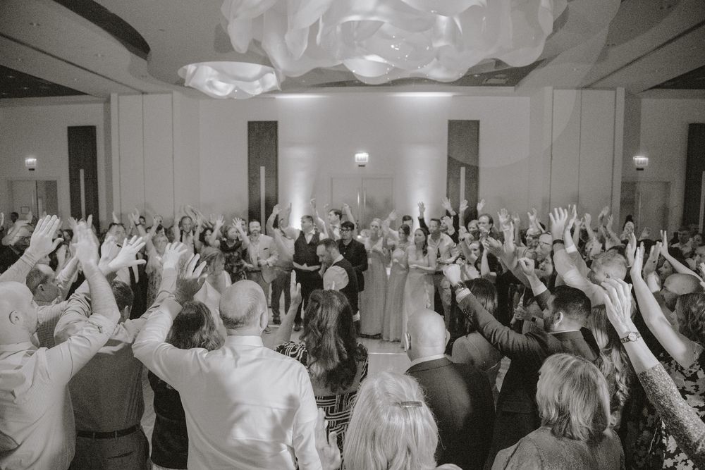 Joyful wedding reception crowd raising hands in a stunning ballroom during a celebratory dance moment.