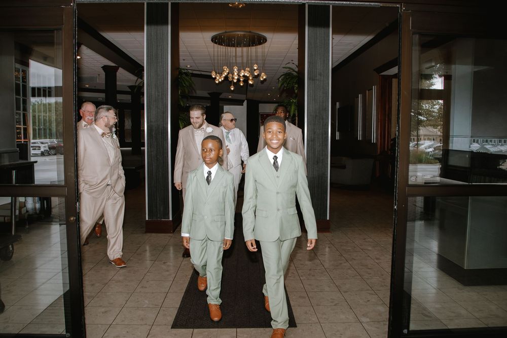 Groomsmen in sage green suits walking through elegant venue entrance during wedding ceremony.
