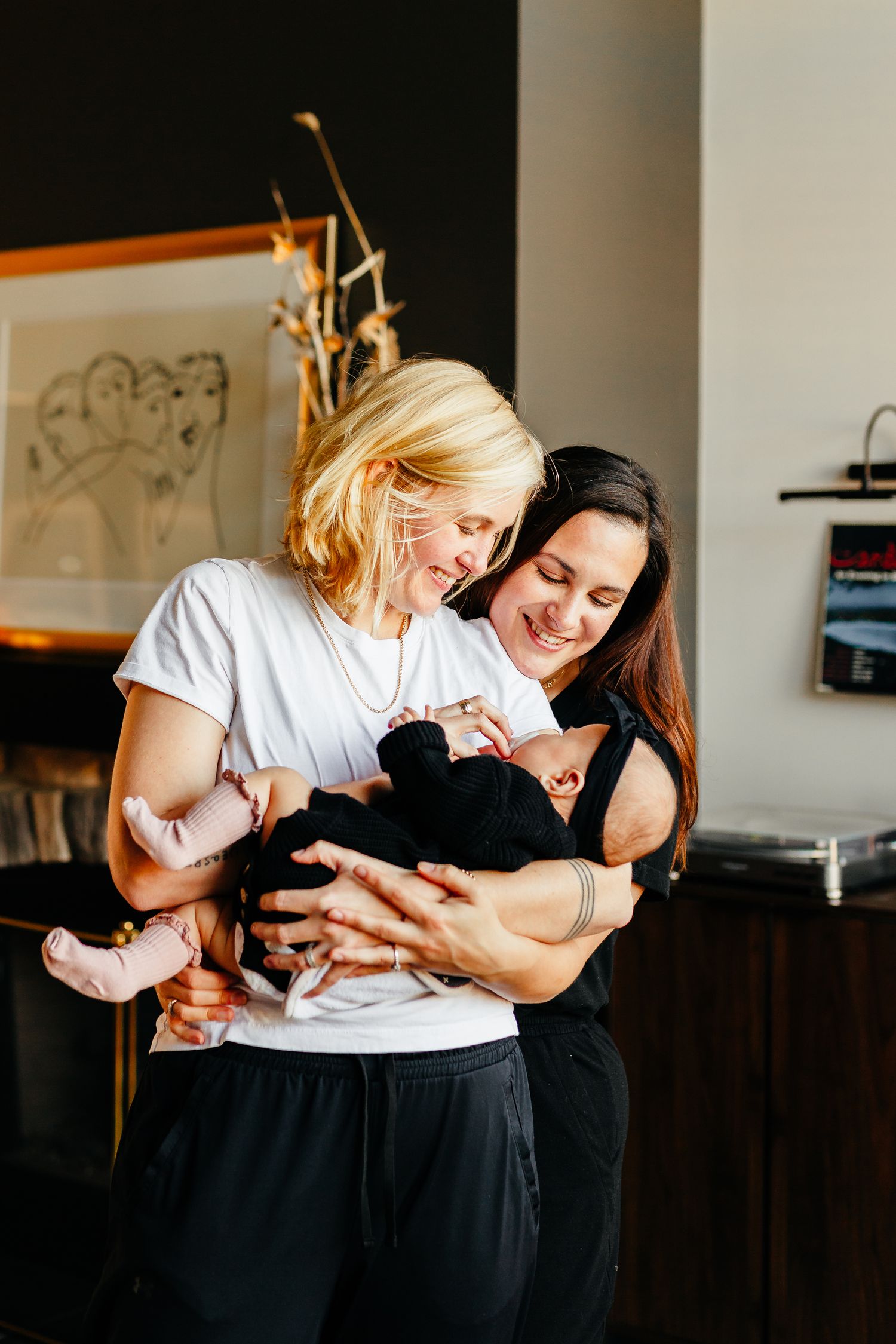 Two smiling women lovingly hold and cuddle a newborn baby in a bright, stylish home with framed artwork on the wall.