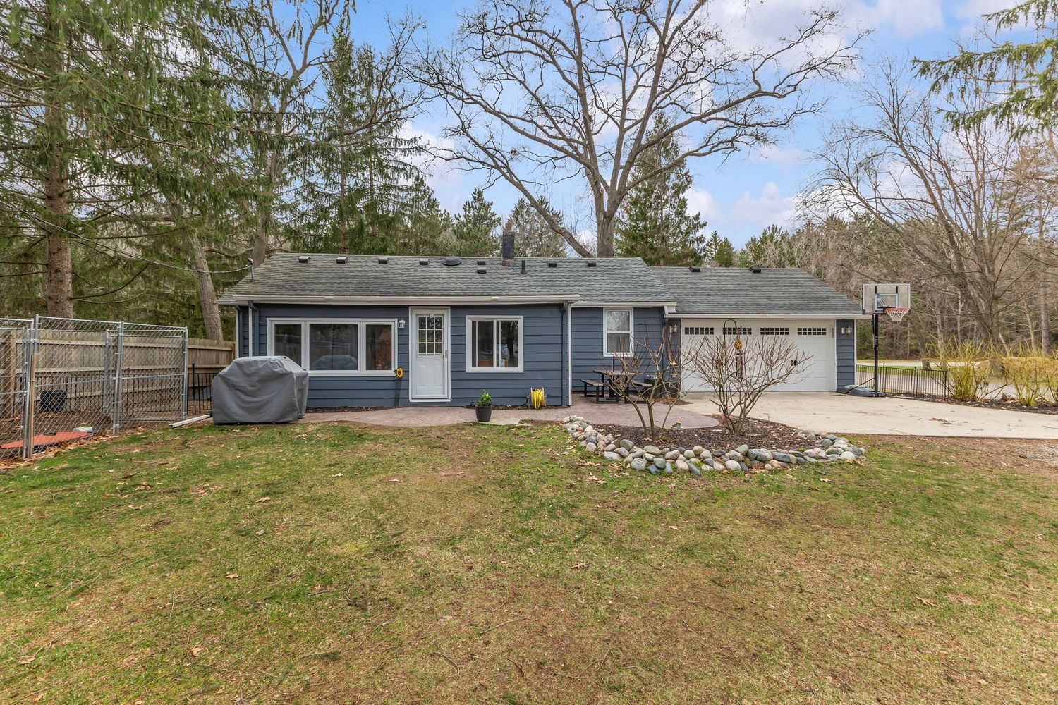 Blue ranch-style home with attached garage, green lawn, and mature pine trees in early spring.