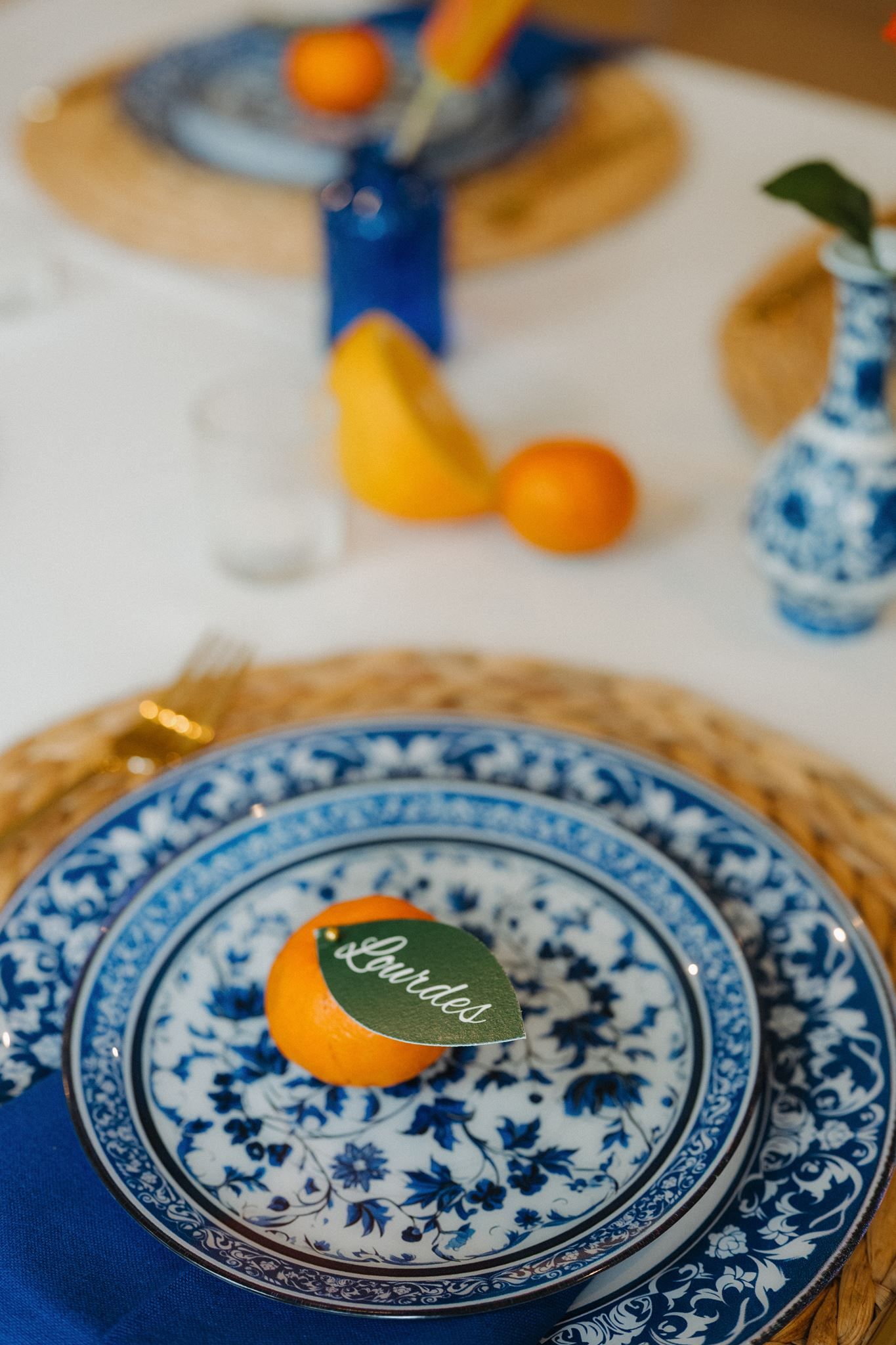 Blue and white floral plates with orange place card on woven charger, blue napkin, citrus fruits, and Delft vases.
