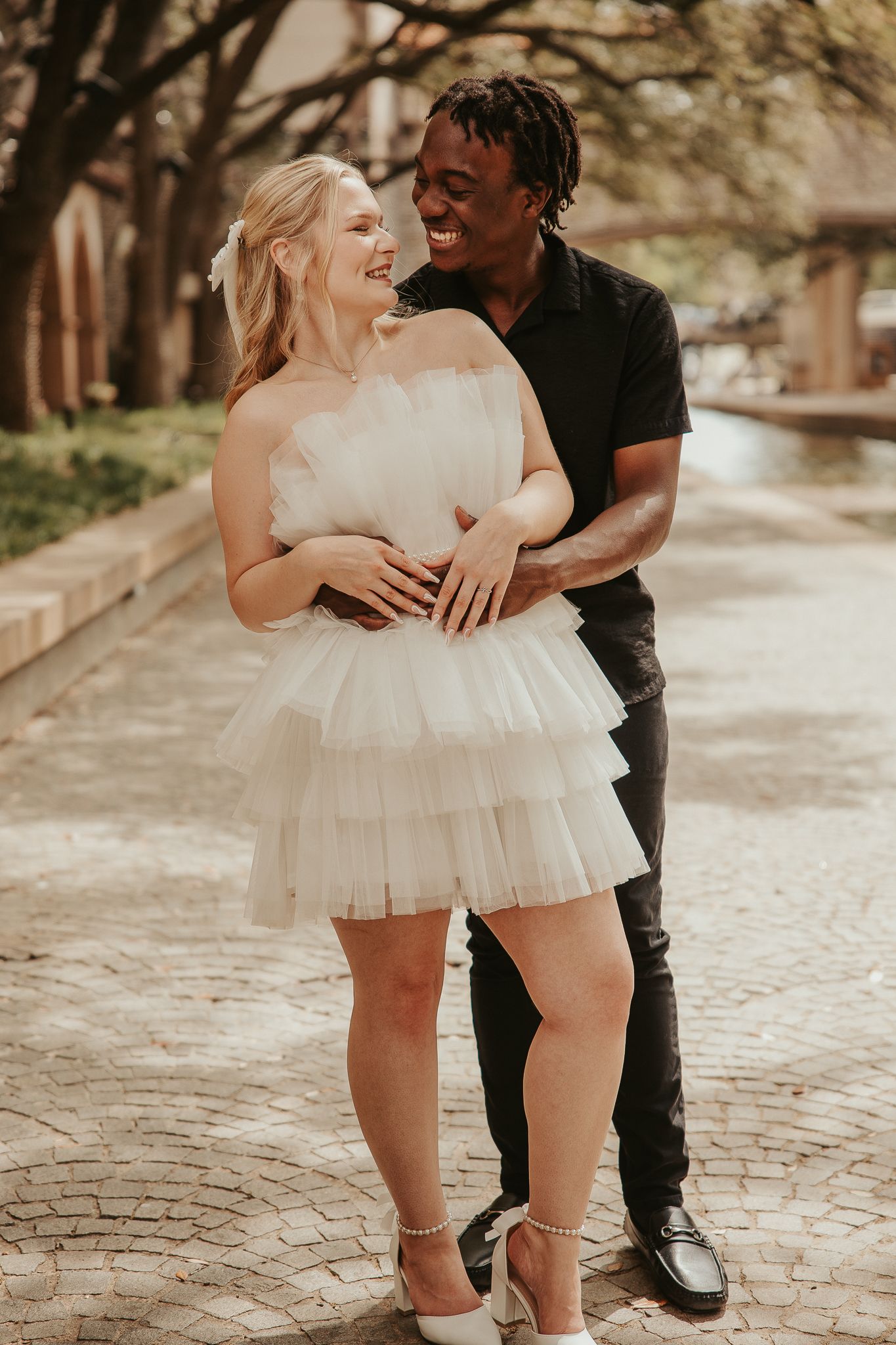A joyful couple poses outdoors, woman in white tulle mini dress and heels, man in black outfit, embracing on brick path.