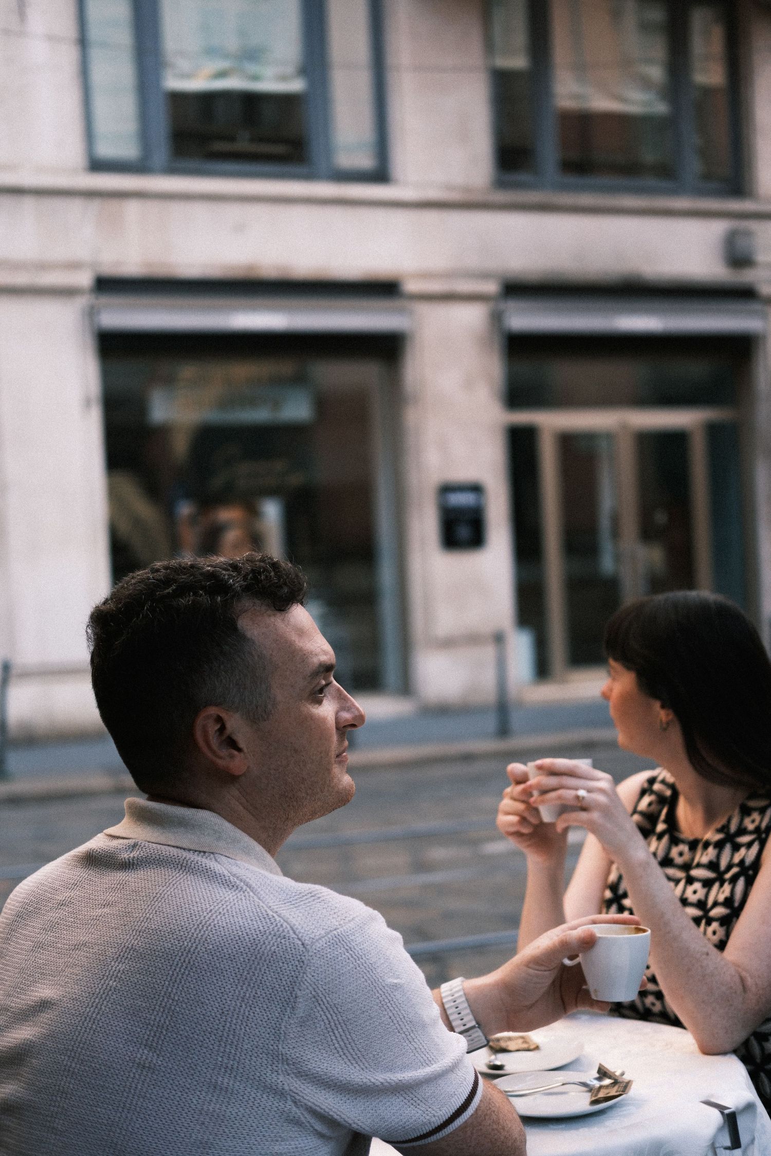 Couple enjoying coffee at an outdoor European cafe table, man in polo shirt conversing with woman in patterned dress.