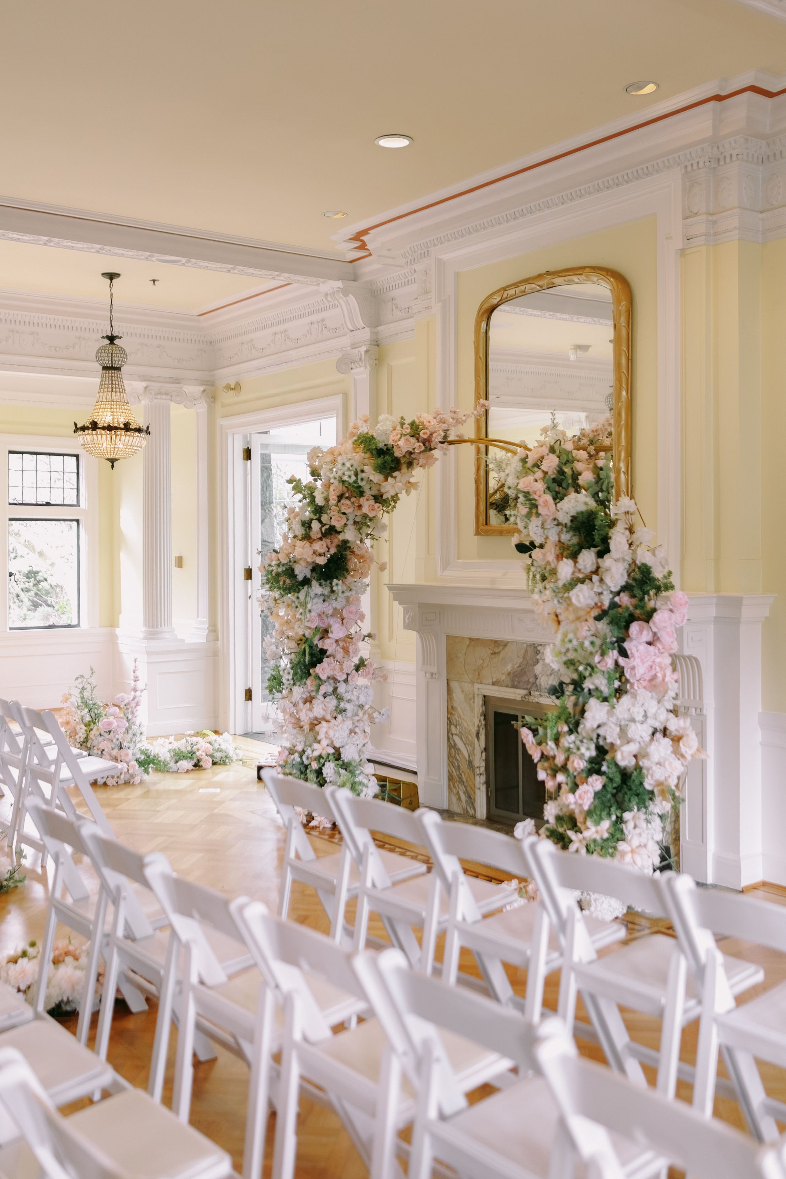 Elegant wedding ceremony room with white chairs, floral arch of pink blooms near a marble fireplace, and gold mirror.