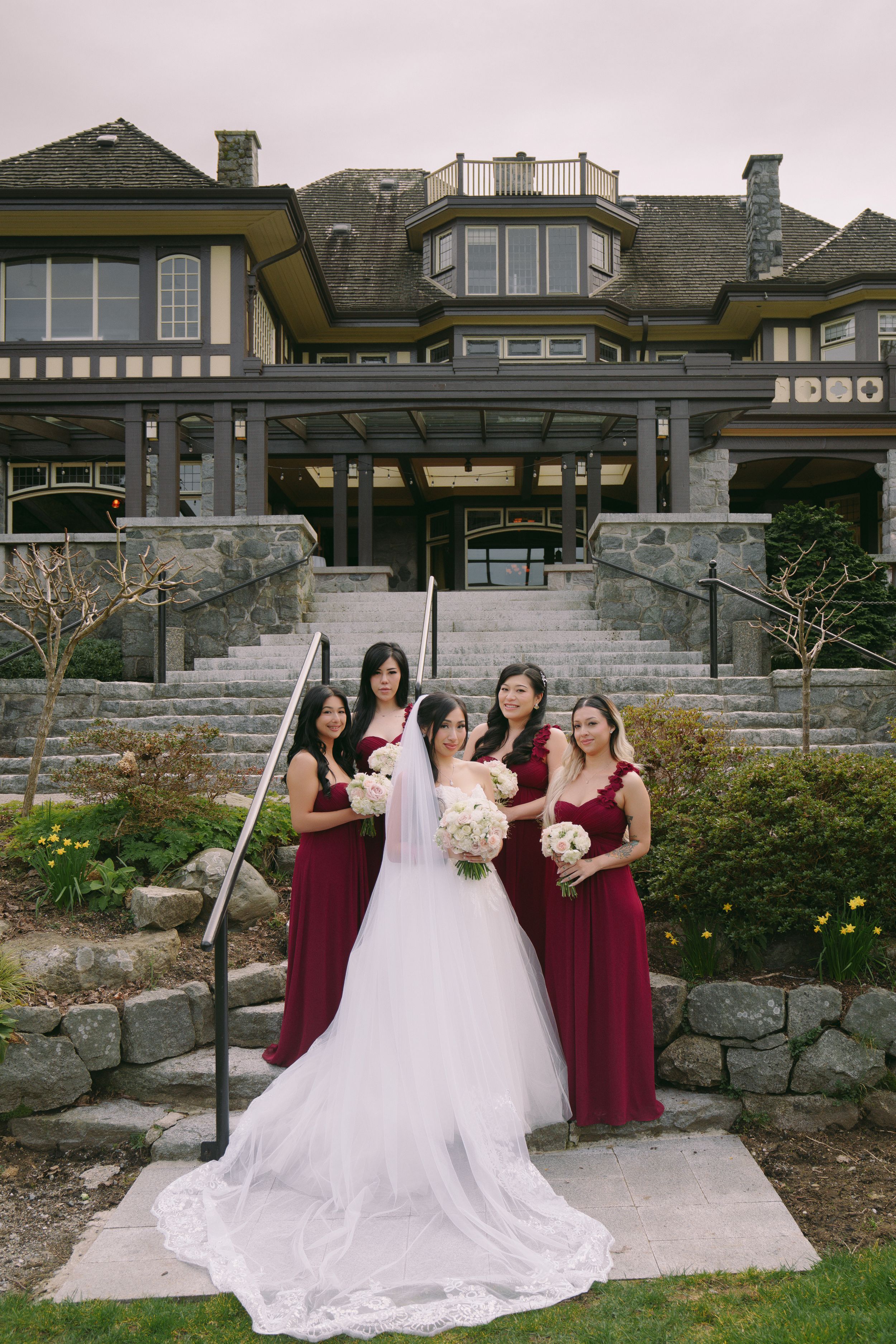 Bride in white gown with bridesmaids in burgundy dresses holding bouquets outside elegant stone mansion.