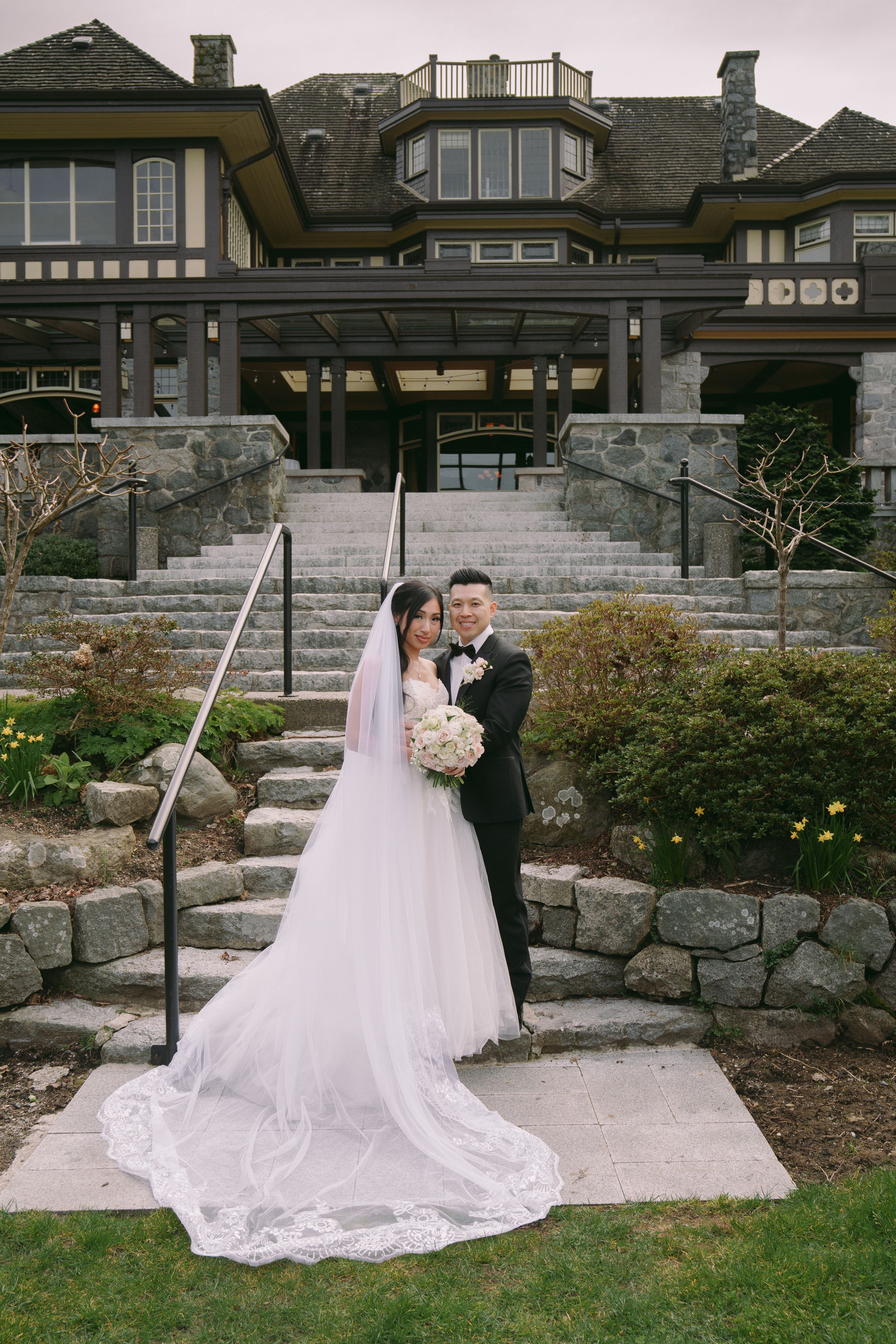 Bride and groom pose on stone steps outside an elegant manor, bride wearing a long lace veil and holding a white bouquet.