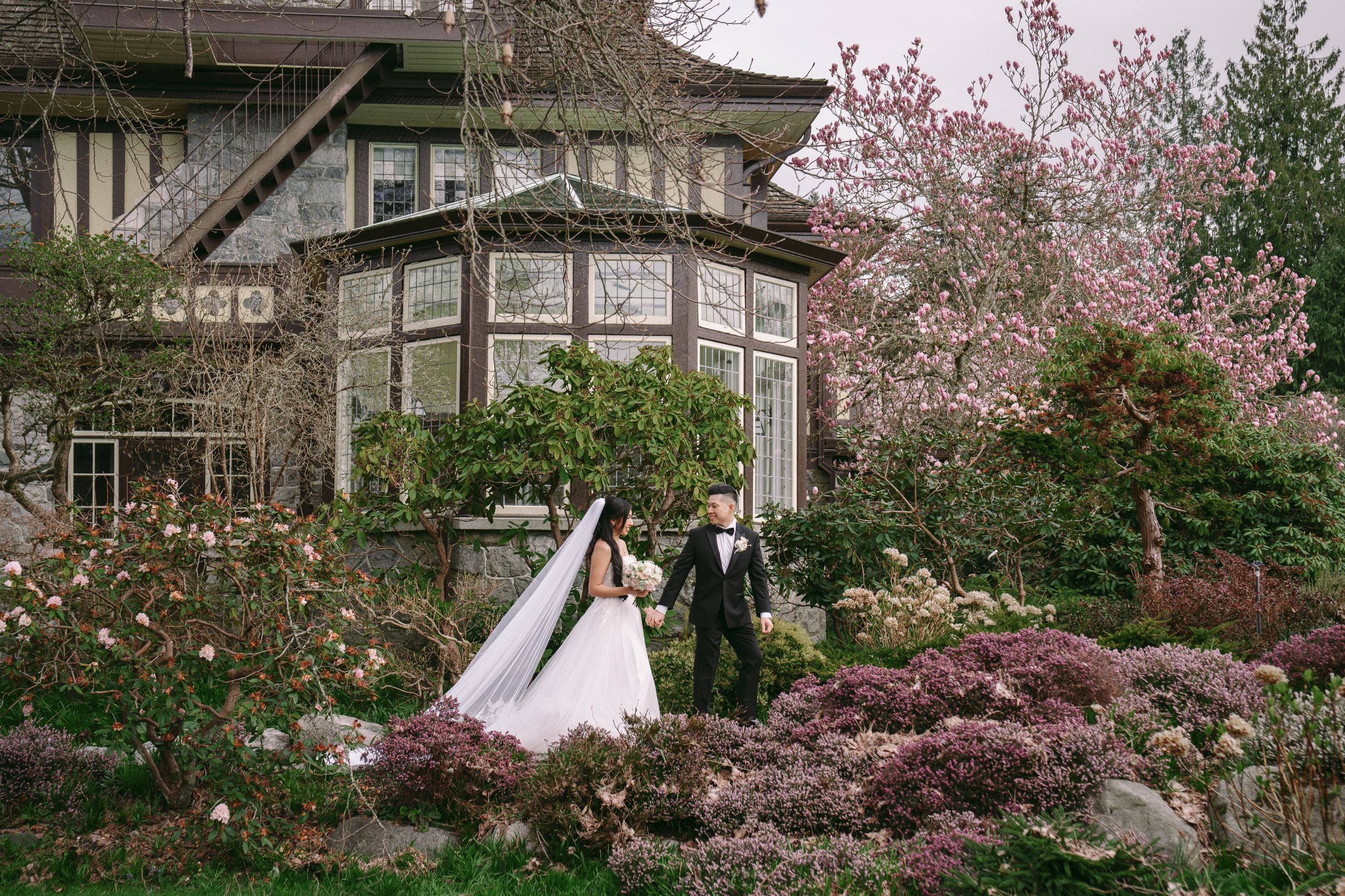 Bride and groom walk hand in hand through a lush garden with blooming cherry blossoms beside an elegant manor.