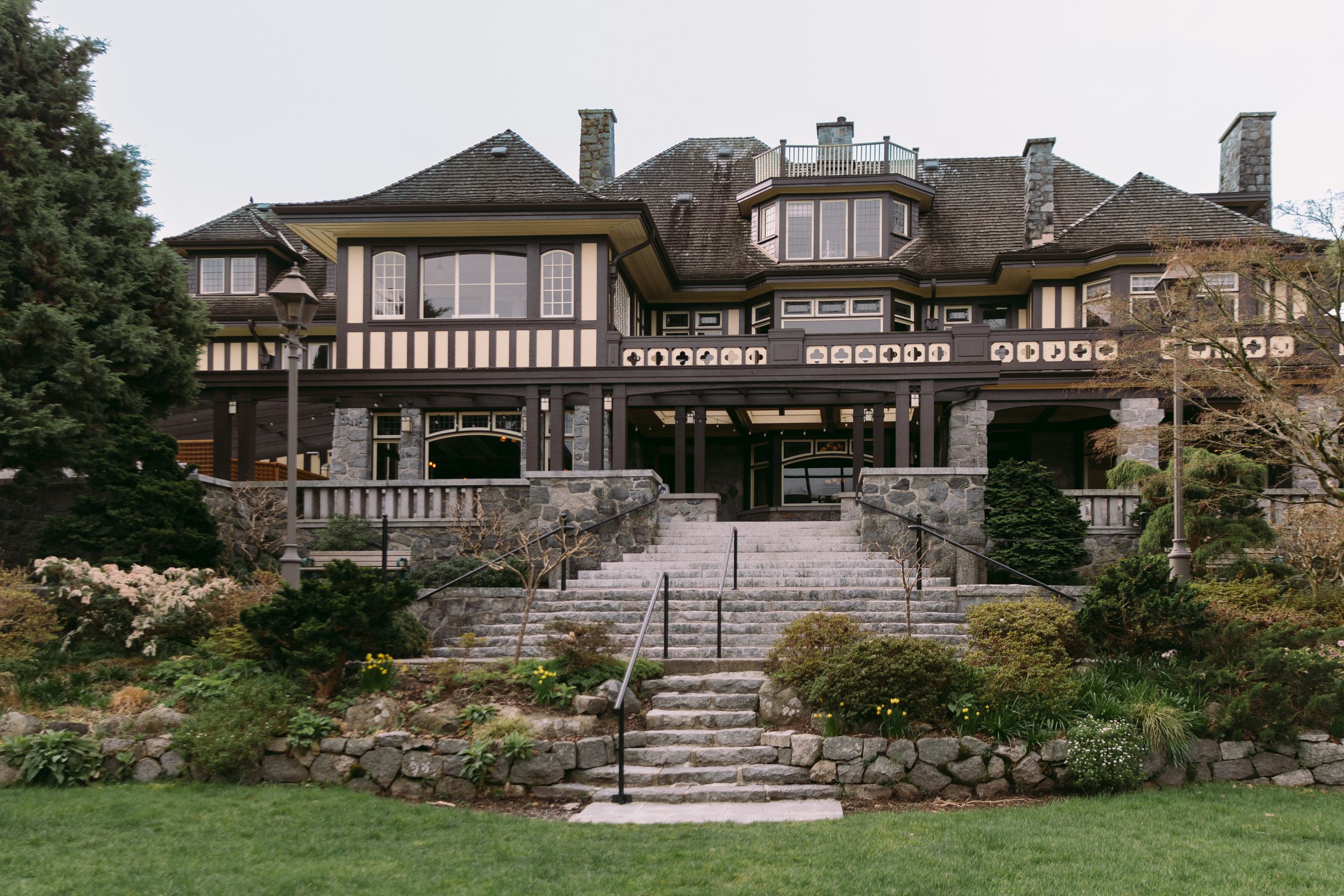 Grand Tudor-style mansion with stone steps, manicured gardens, and elegant architecture surrounded by lush greenery.
