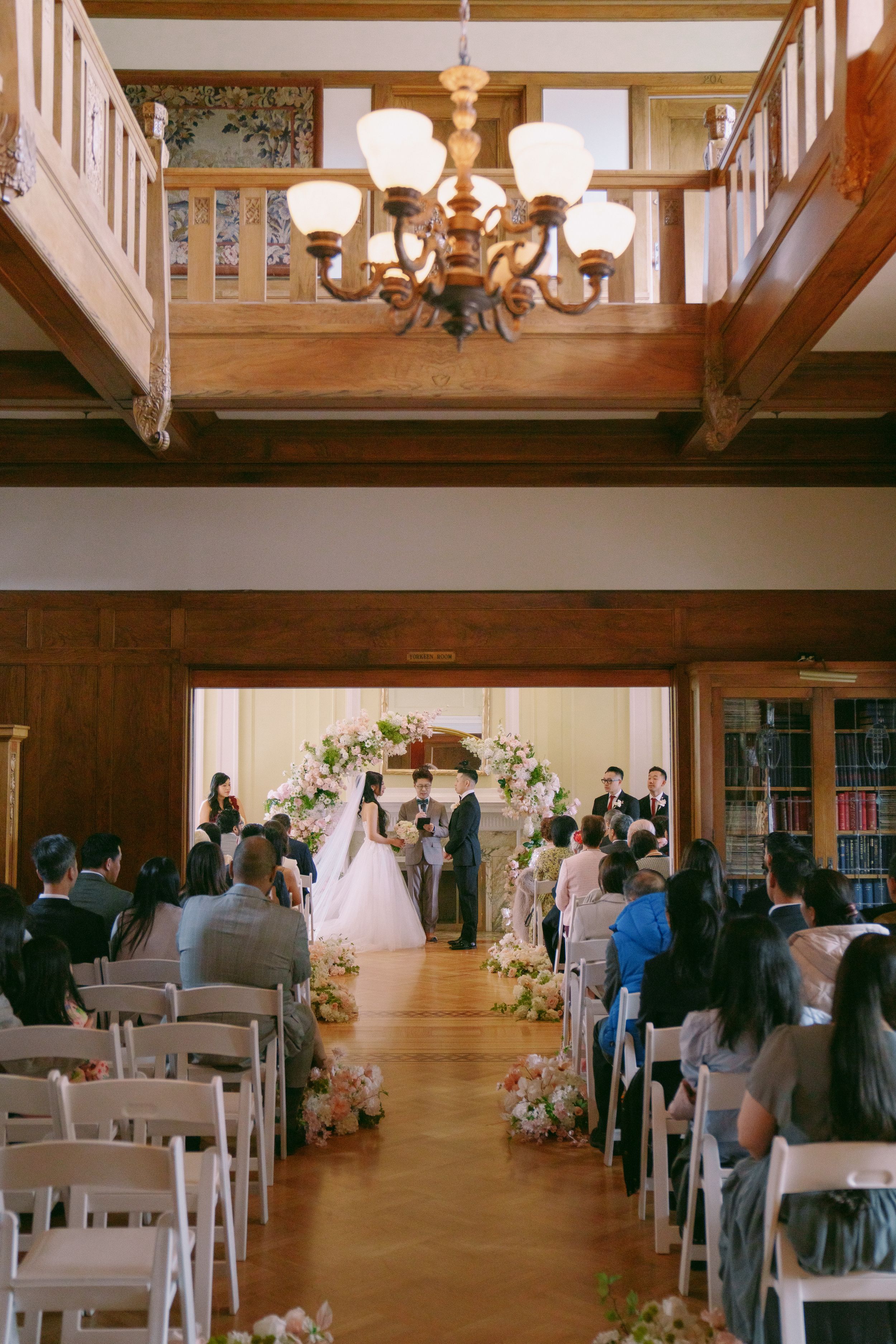 Indoor wedding ceremony in elegant wood-paneled room with floral arch, white chairs, and chandelier overhead.