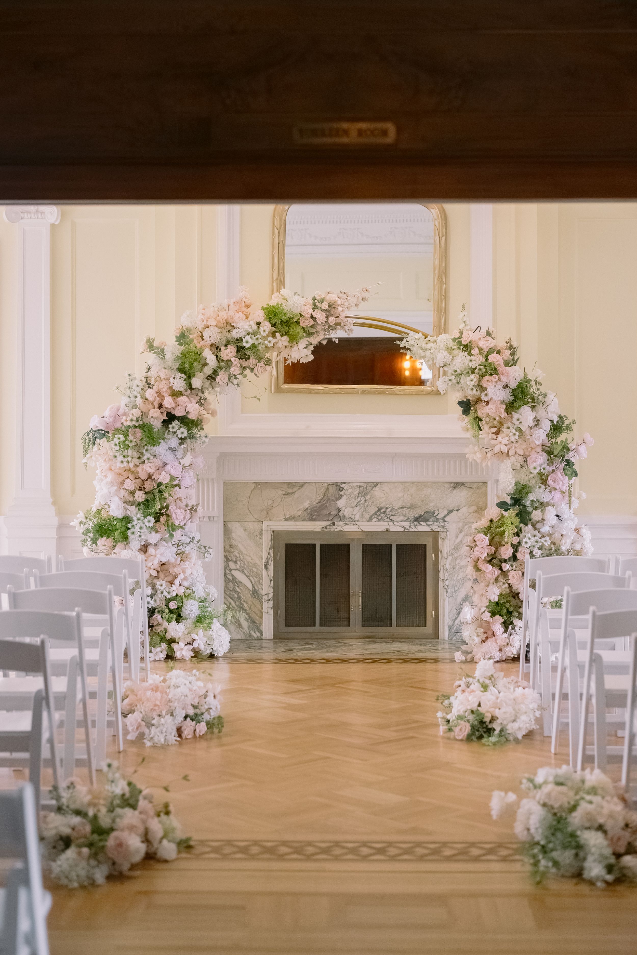 Elegant wedding ceremony arch adorned with blush pink and white flowers beside a marble fireplace in a bright ballroom.
