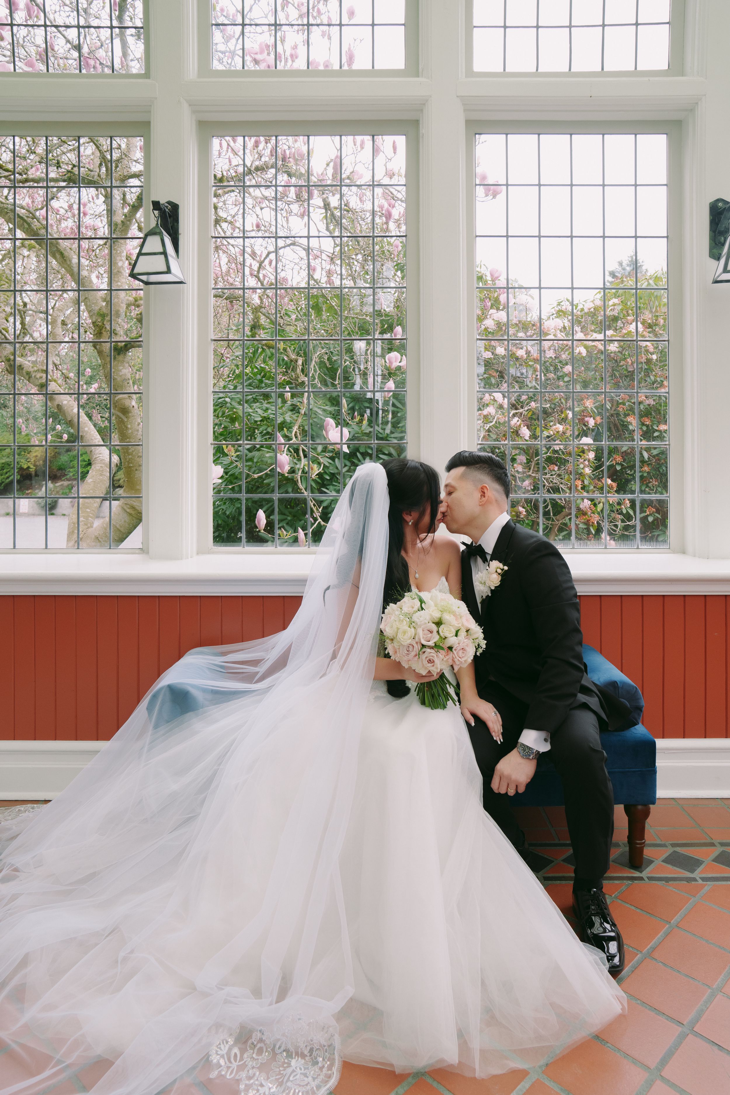 Bride and groom share a romantic kiss by large windows with blooming spring trees, bride holding white bouquet in elegant gown.