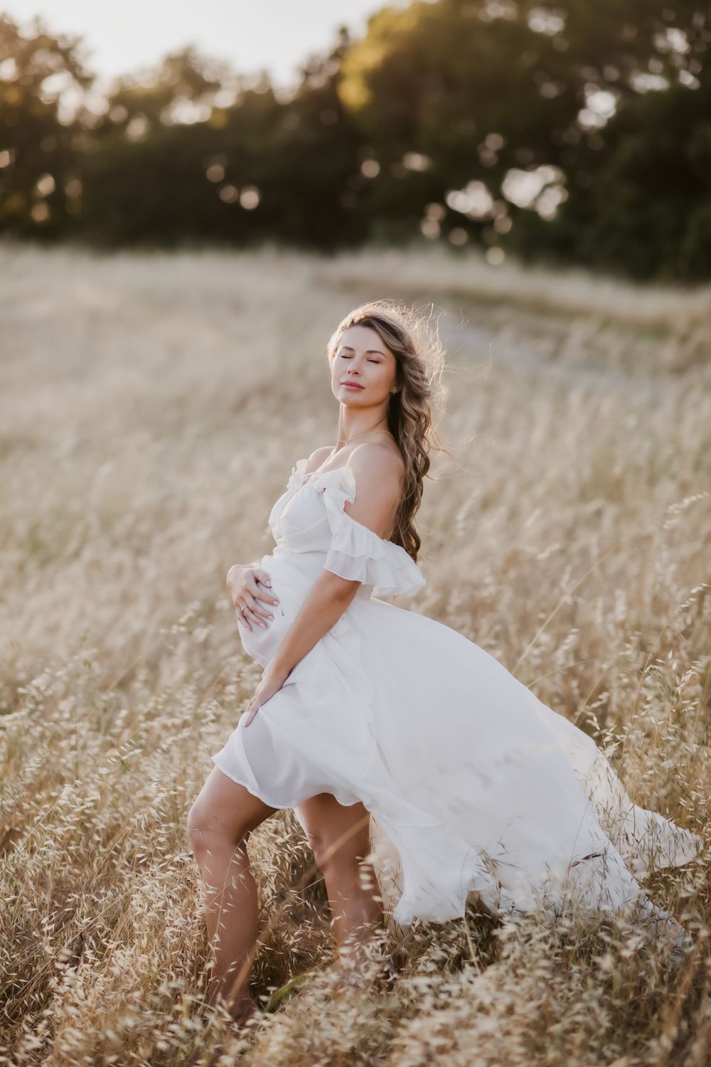 Expectant mother in white flowy gown poses in golden field for maternity photos. Exeter Andover Boxford maternity photographer