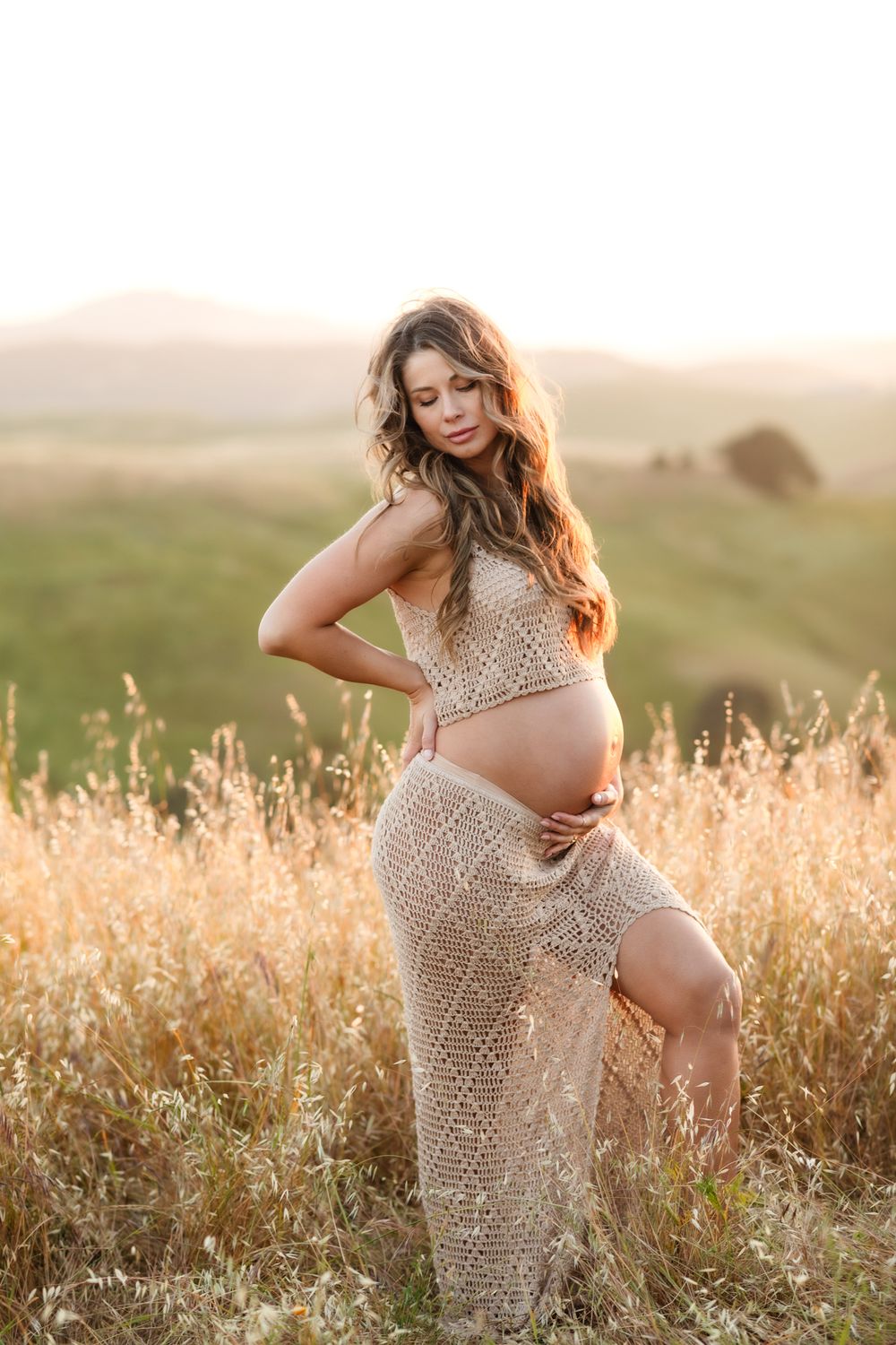 Expectant mother in tan crochet outfit poses in golden field with hills in background. Chelmsford Alton Bay maternity photographer